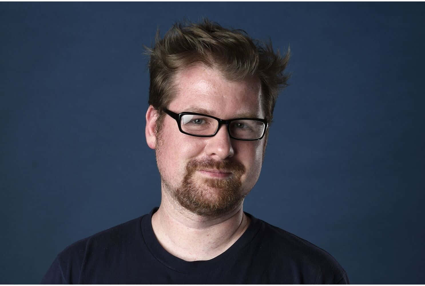 Justin Roiland 桌布 壁紙 — 一位戴眼鏡留鬍鬚的男子在純深藍色攝影棚背景前溫柔微笑。