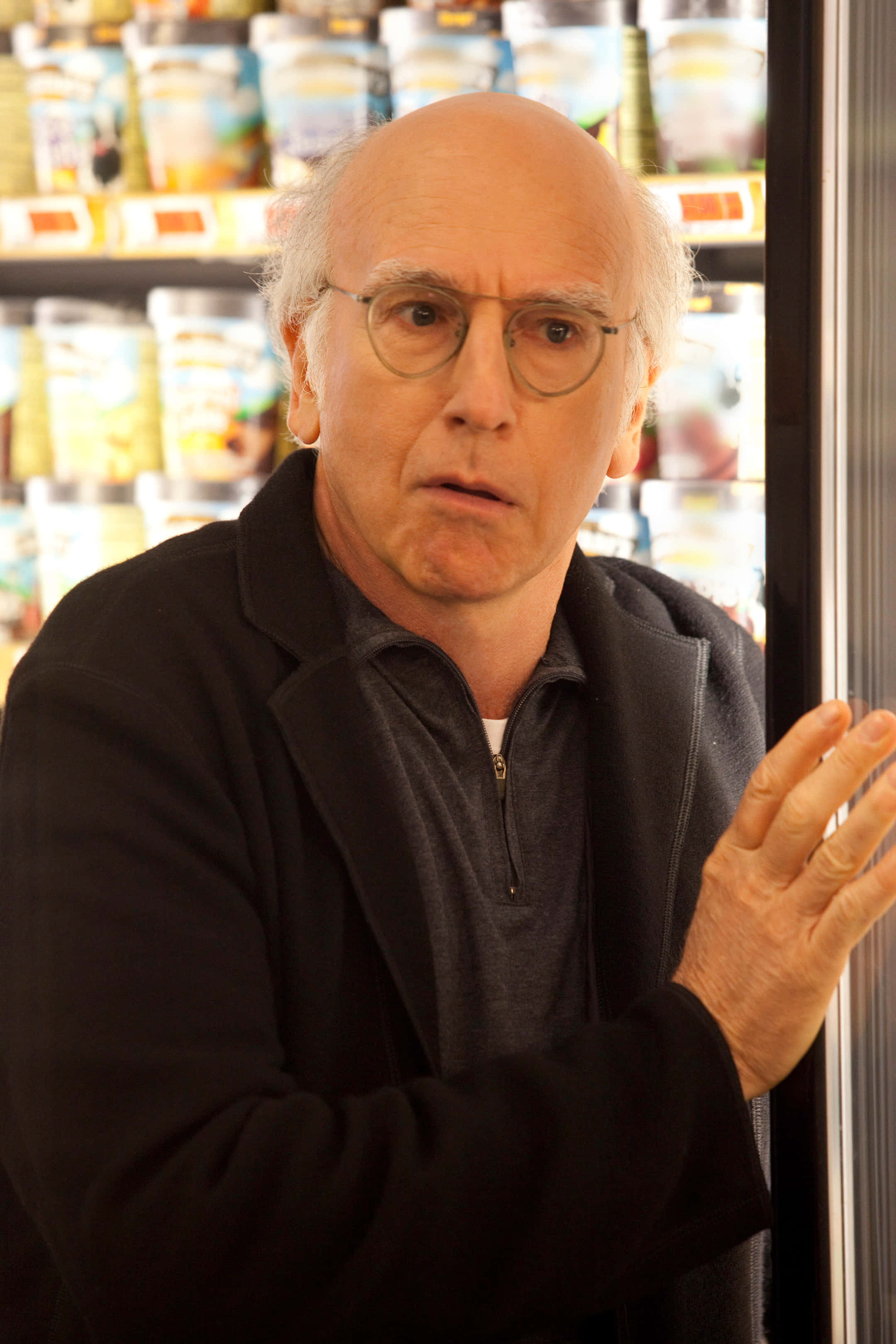 larry david 桌布 壁紙 — 一位戴眼鏡的光頭男子站在雜貨店通道中，表情驚訝。