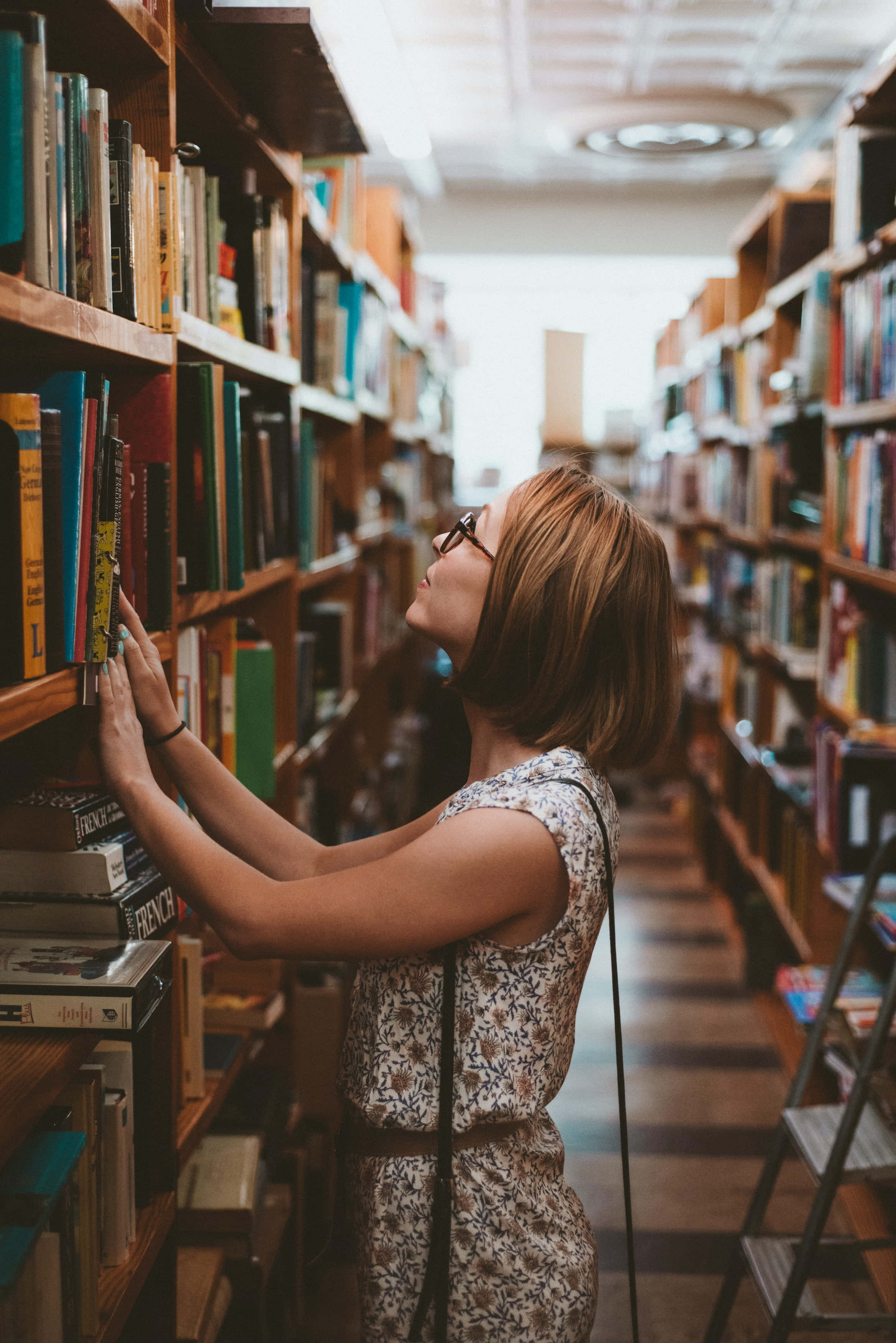 學習 桌布 壁紙 — 一位戴眼鏡的女性在燈光溫暖的圖書館走廊中，伸手去拿木書架上高處的書。