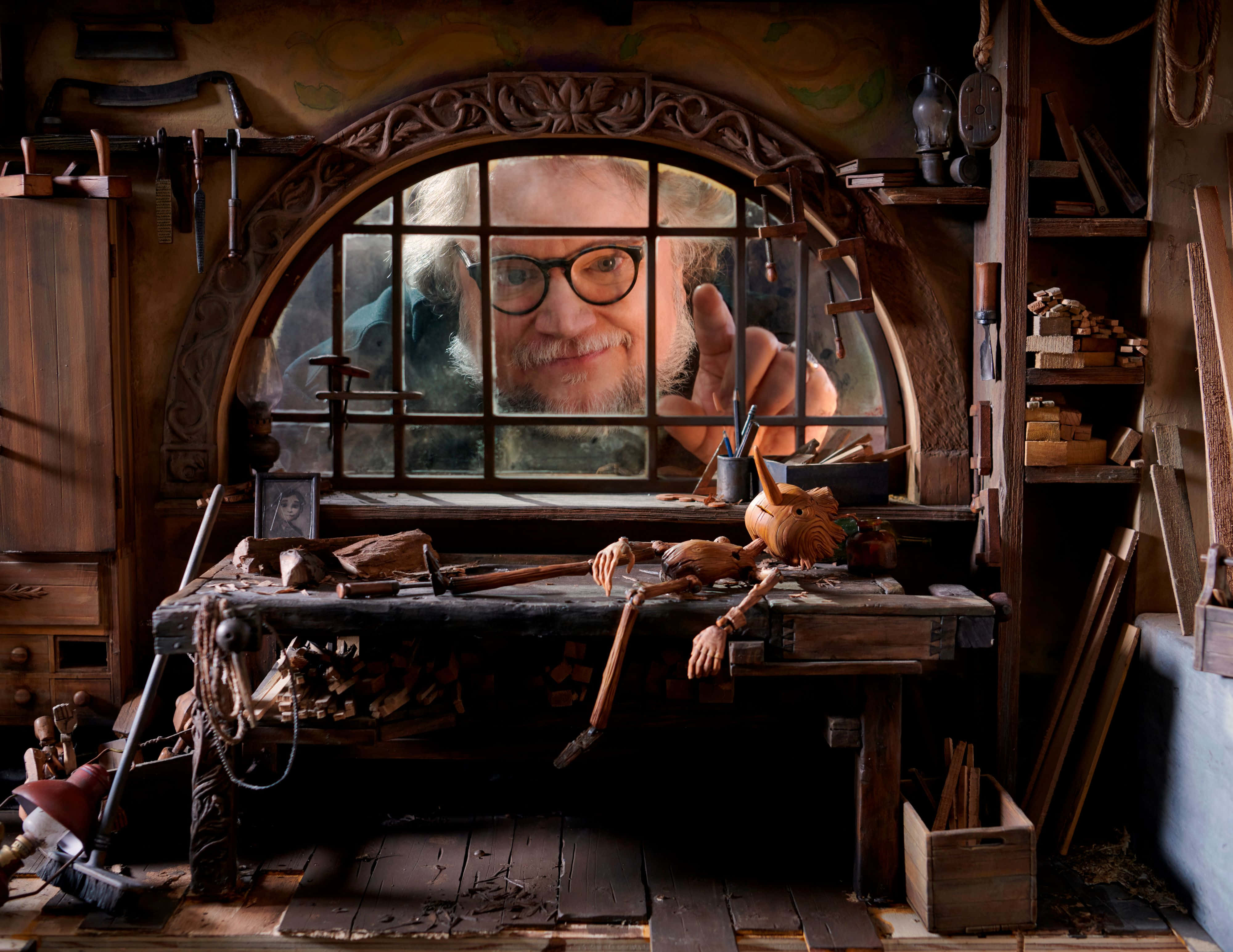 Guillermo Del Toro's Pinocchio 桌布 壁紙 — 一位戴眼鏡的工匠透過木製拱形窗戶溫暖地凝視著他雜亂的工作室，裡面充滿了工具和木雕。