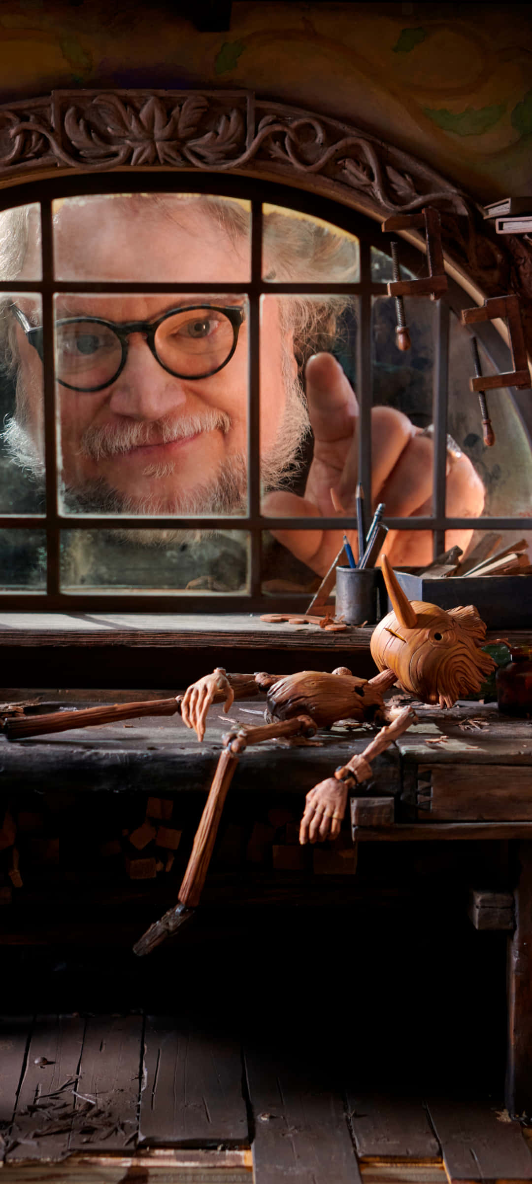 Guillermo Del Toro's Pinocchio 桌布 壁紙 — 一位戴眼鏡的微笑工匠透過木窗看著躺在雜亂工作台上的木偶。