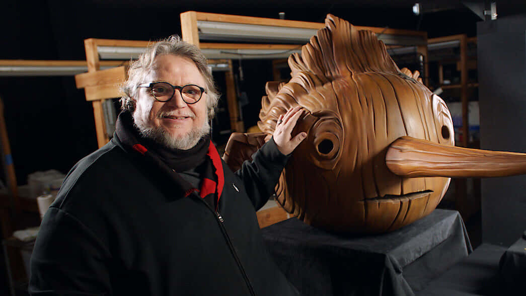 Guillermo Del Toro's Pinocchio 桌布 壁紙 — 一位戴眼鏡的微笑男子在工作室環境中輕輕觸摸一個大型精雕細琢的木魚雕塑。