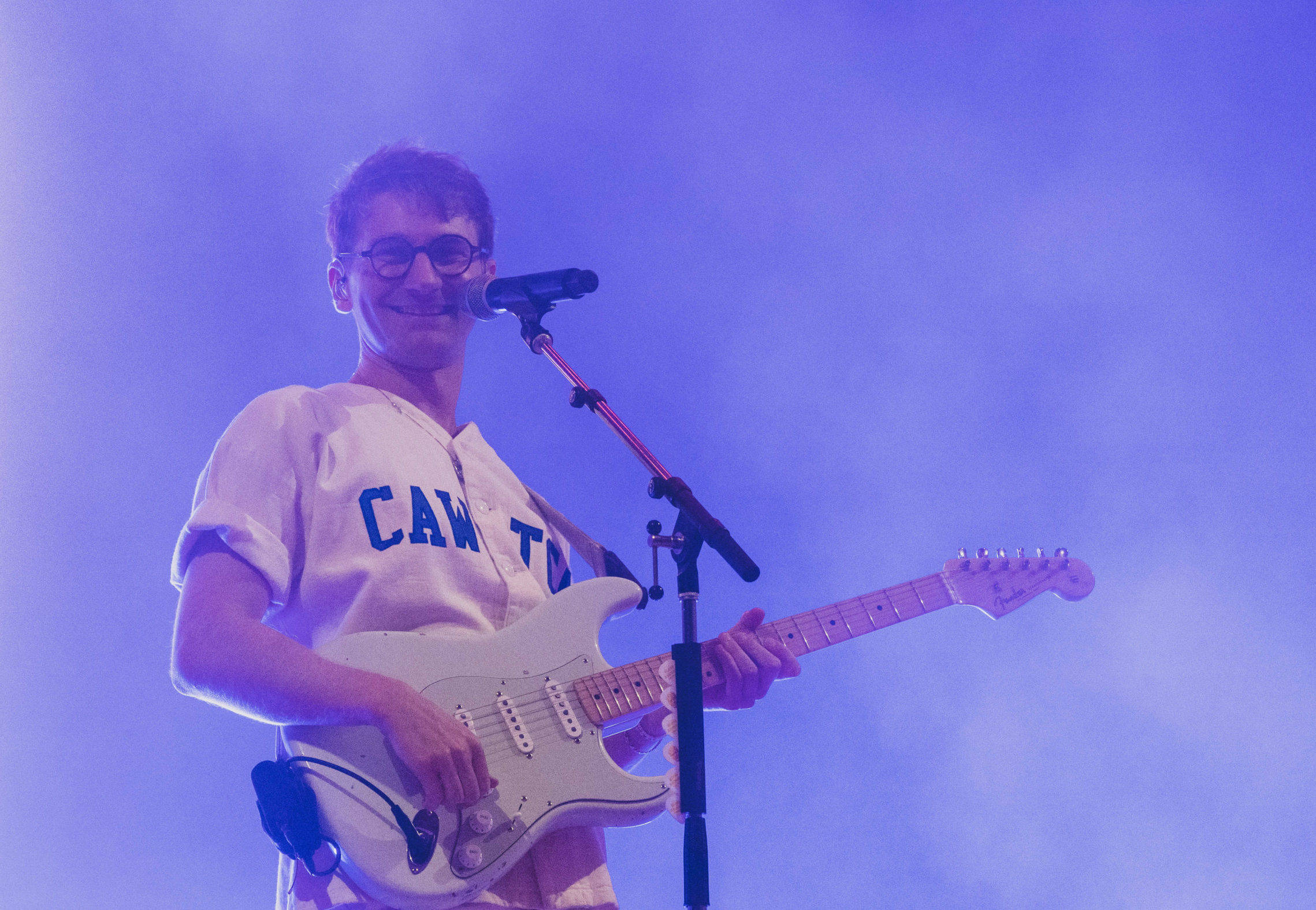Glass Animals Bayley 壁紙 — 一位戴眼鏡的微笑音樂家在紫色舞台燈光和煙霧中彈奏白色電結他。