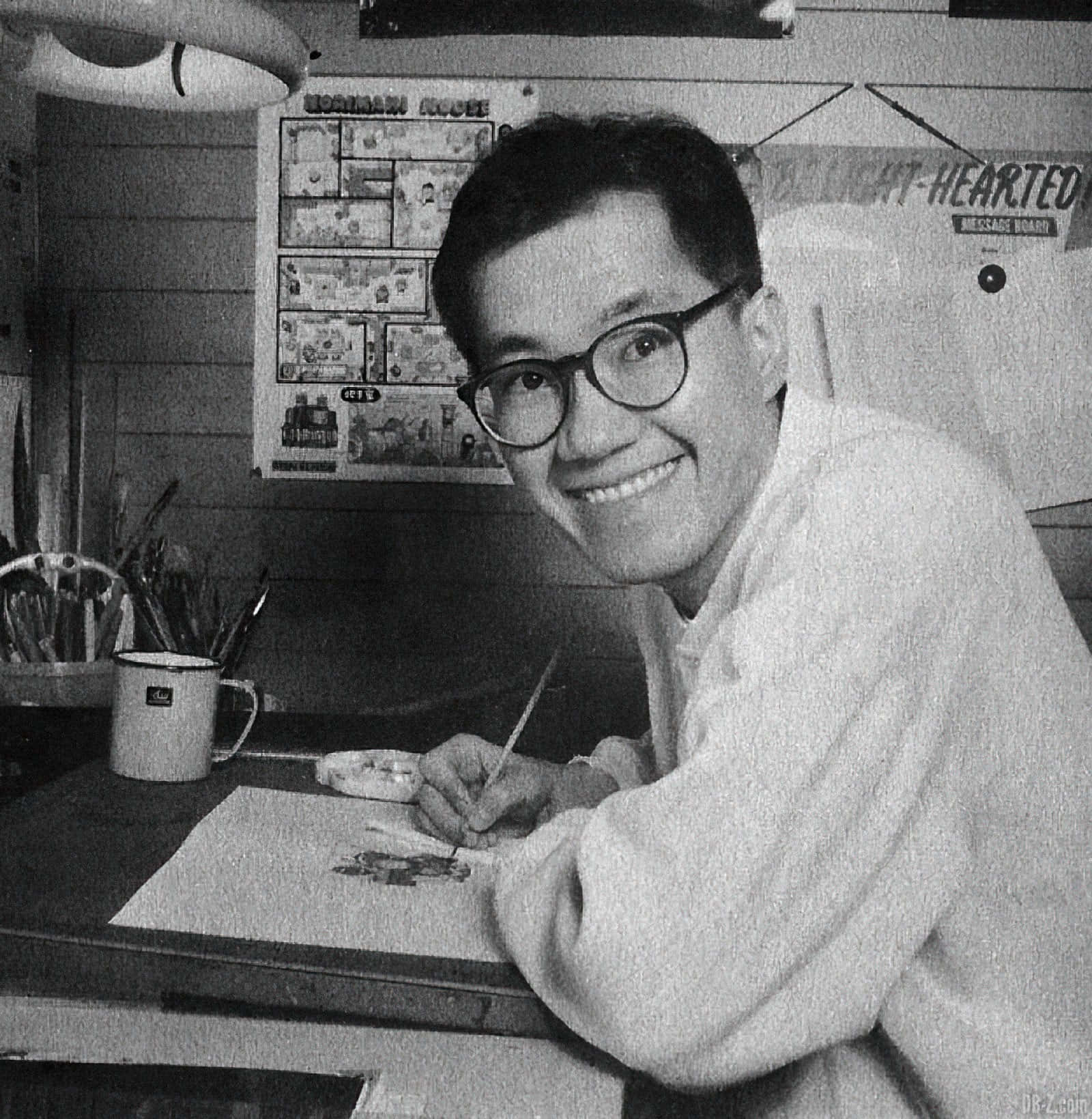 Akira Toriyama 桌布 壁紙 — 一位戴眼鏡的快樂藝術家微笑著在雜亂的桌子上畫畫，牆上貼滿漫畫海報。