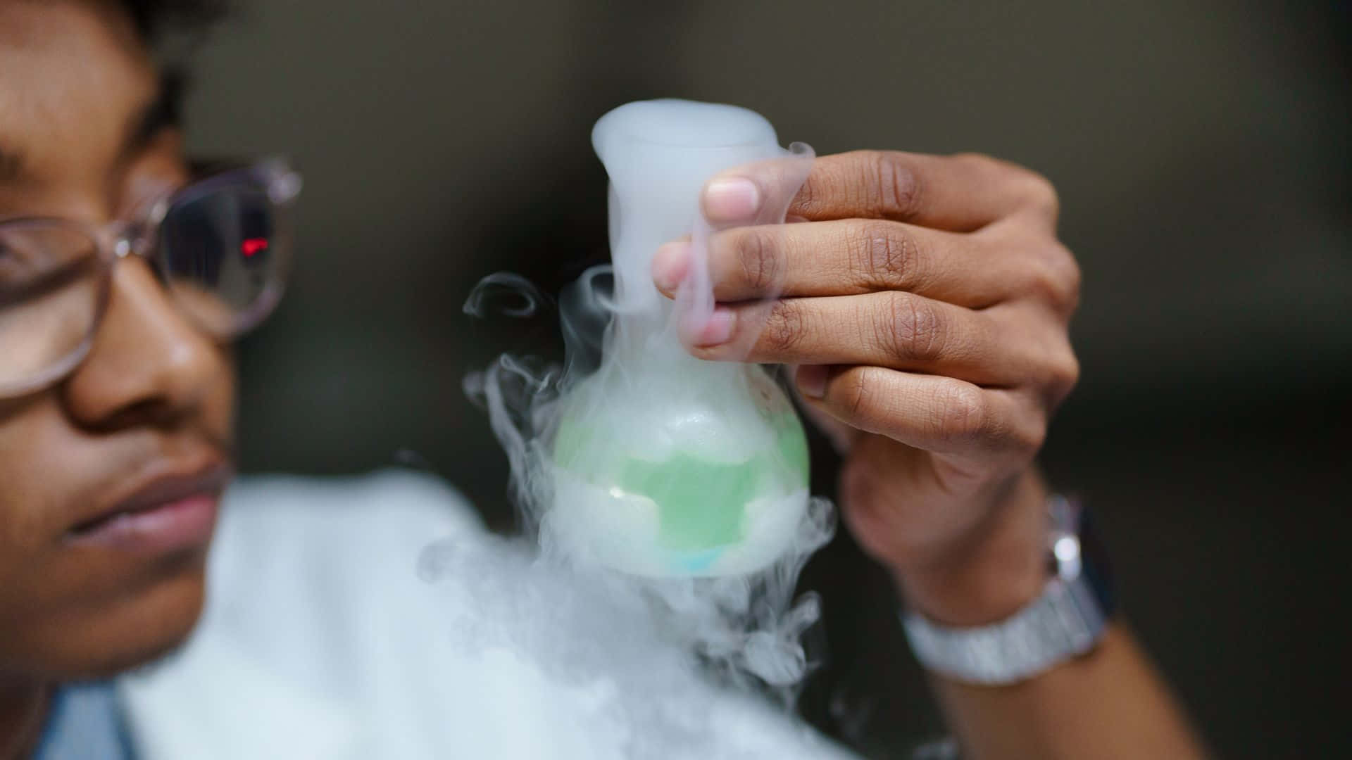 Green Chemistry 桌布 壁紙 — 一位戴眼鏡的科學家在昏暗的實驗室中手持一個冒煙的燒瓶，裡面裝有發光的綠色液體。