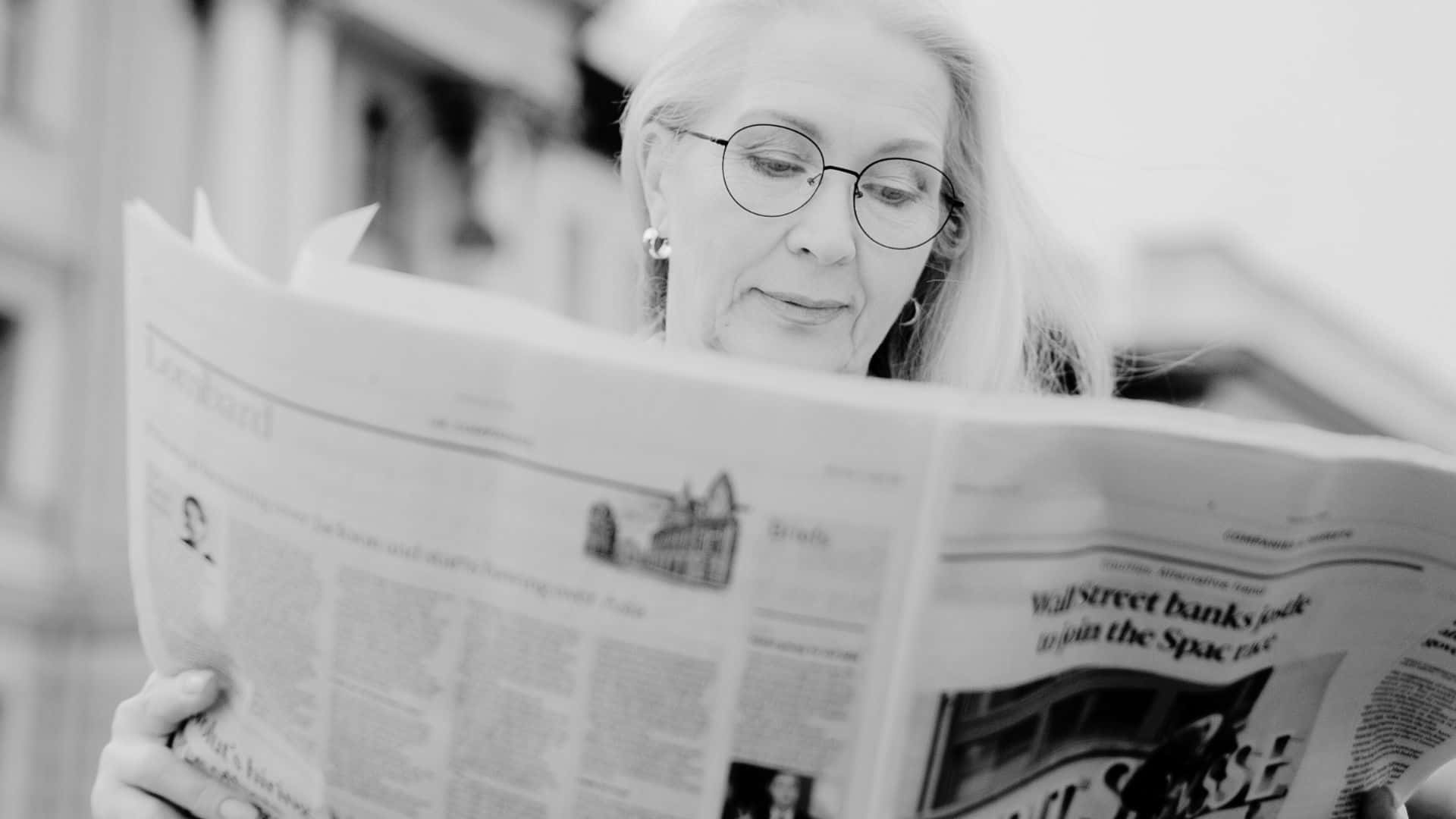 Black And White Newspaper 桌布 壁紙 — 一位戴眼鏡的老年女性在戶外閱讀報紙，黑白影像，背景是模糊的城市景觀。