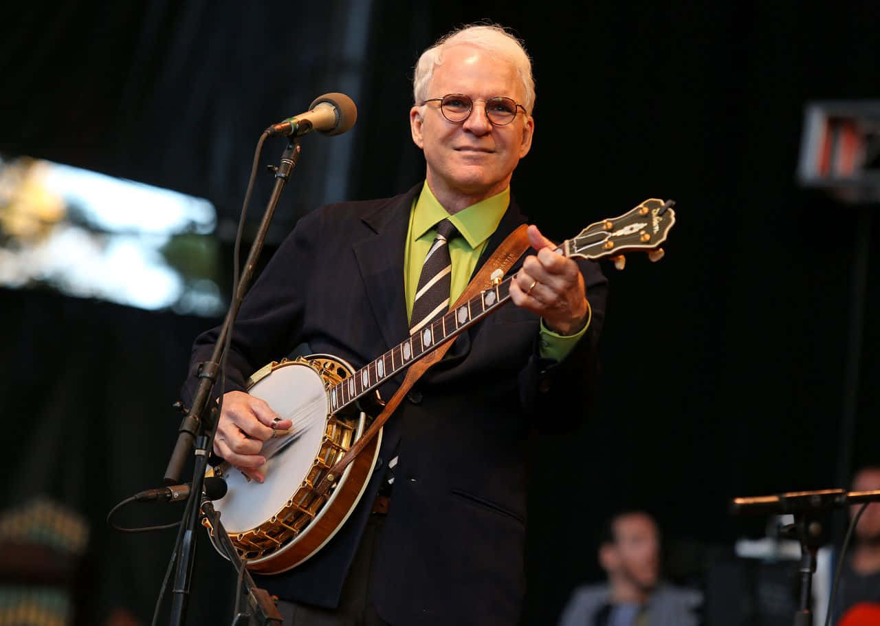 steve martin 桌布 壁紙 — 一位戴眼鏡的長者，在昏暗的舞台上，於聚光燈下微笑著彈奏班卓琴。