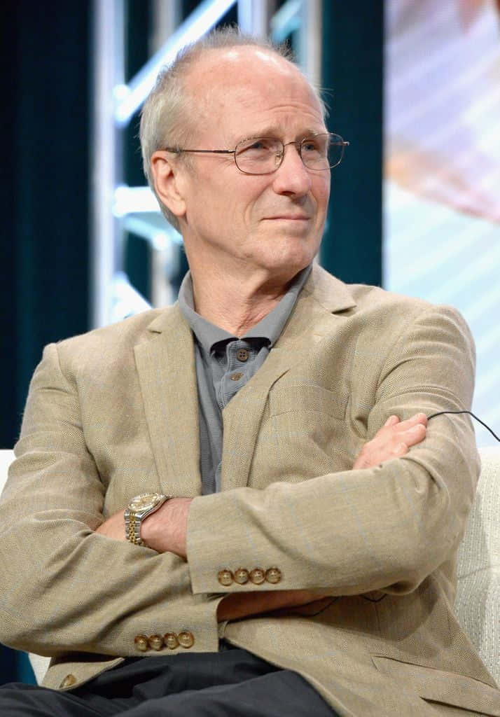 William Hurt 桌布 壁紙 — 一位戴眼鏡的長者冷靜地坐著，雙臂交叉，身穿米色西裝外套，佩戴金色手錶。