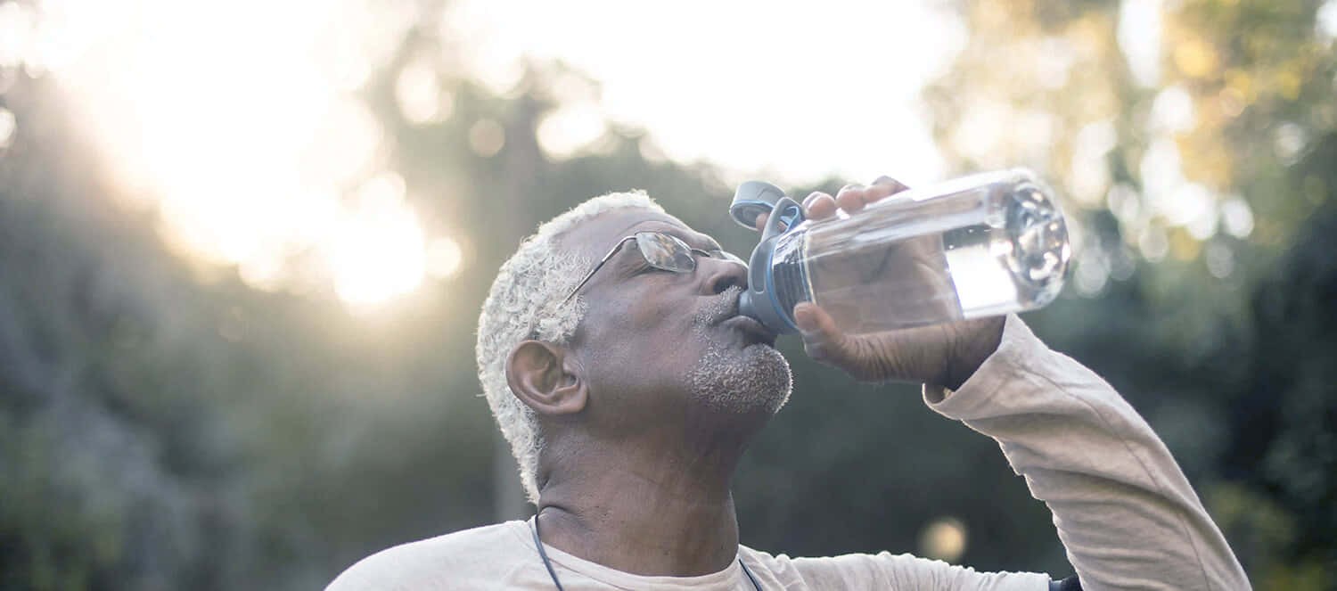 Hydration 桌布 壁紙 — 一位戴眼鏡的長者從透明水樽飲水，背後金色陽光穿過樹木灑落。