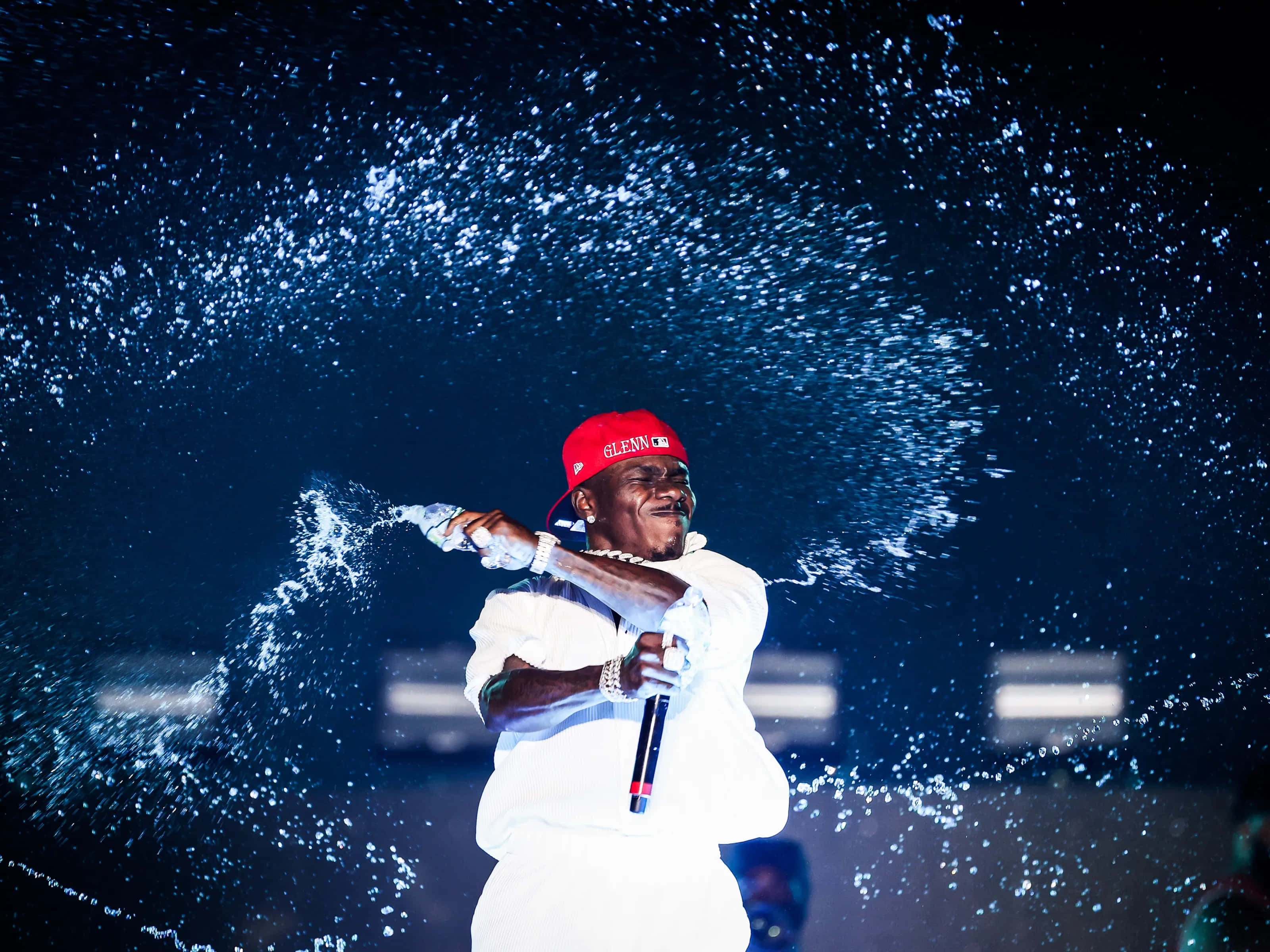Dababy 桌布 壁紙 — 一位戴紅帽、穿白色服裝的表演者在戲劇性的藍色燈光下，在舞台上歡樂地噴灑香檳。