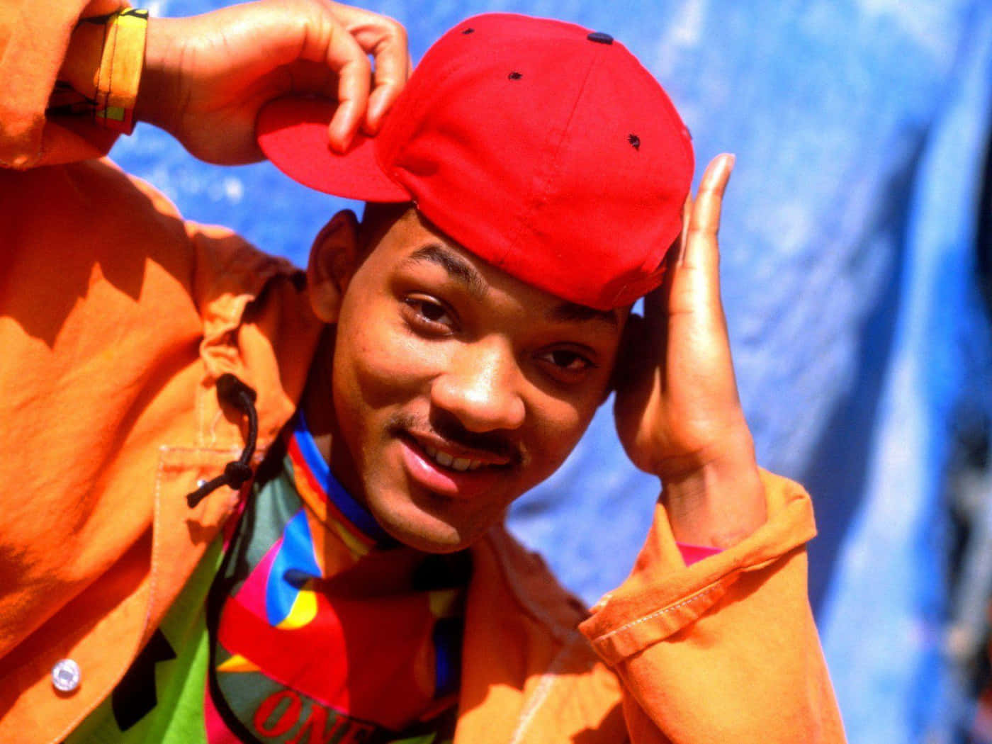 fresh prince 桌布 壁紙 — 一位戴紅色帽子和橙色外套的年輕男子在鮮豔的藍色背景下微笑著調整帽子。