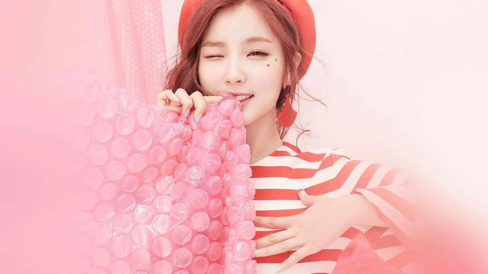 Kpop Kawaii 桌布 壁紙 — 一位戴紅色貝雷帽、穿條紋襯衫的微笑女士，在柔和的粉紅色背景前俏皮地拿著粉紅色泡泡紙。