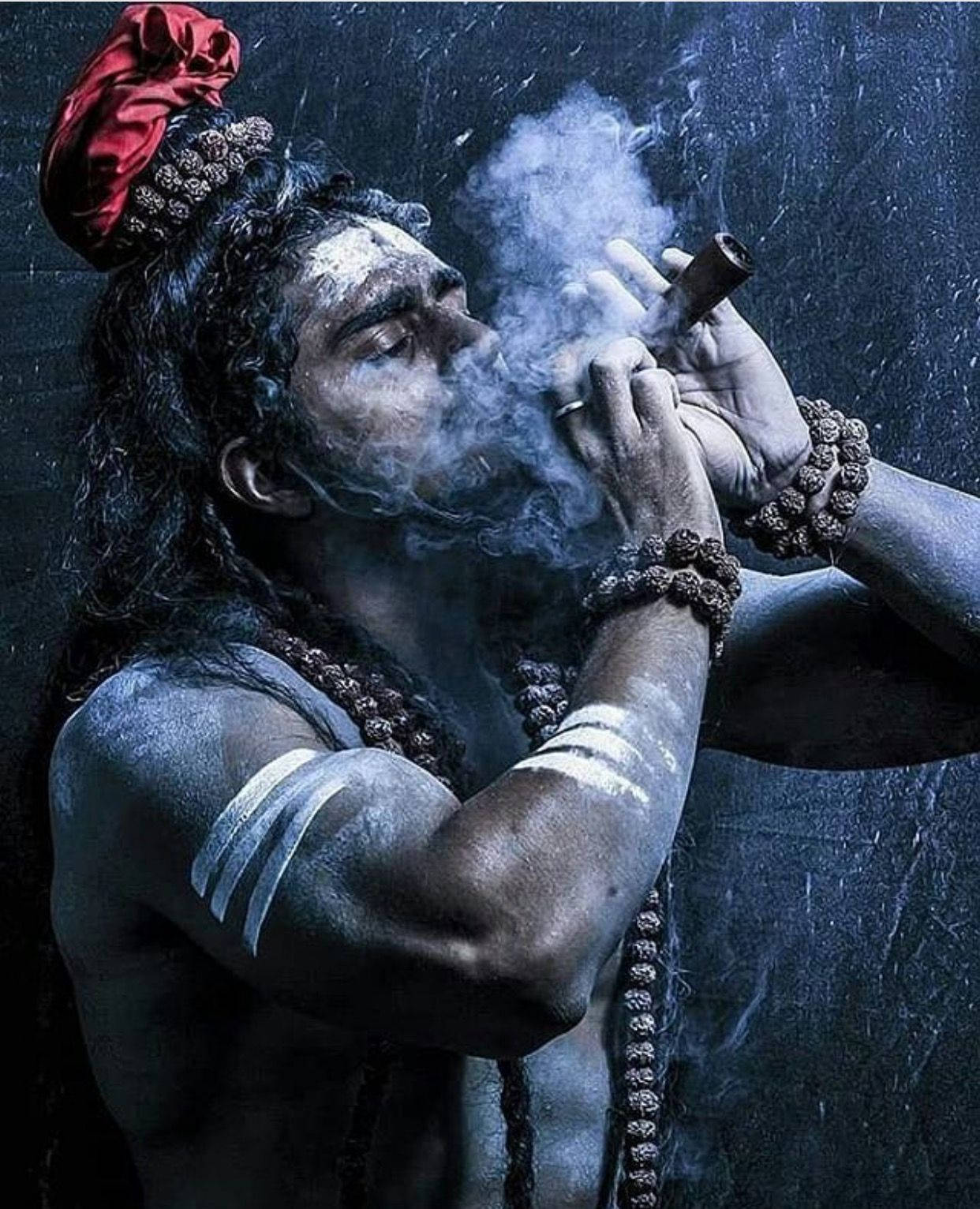 hindu iPhone 桌布 壁紙 — 一位戴紅色頭巾、有白色標記的瑜伽士，手持煙斗對著深色紋理牆壁呼出煙霧。