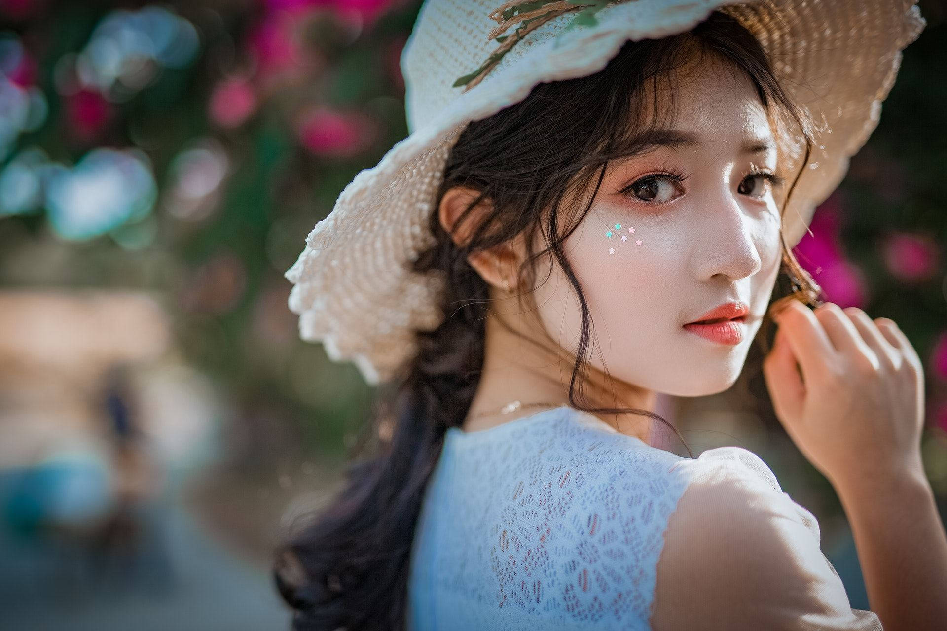 Beautiful Girl 桌布 壁紙 — 一位戴草帽、穿藍色蕾絲上衣的年輕女性，在花卉散景背景下溫柔凝視。
