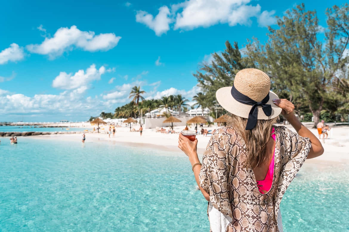 Bahamas Island 桌布 壁紙 — 一位戴草帽嘅女士喺明亮藍天下嘅藍綠色海灘上，喺棕櫚樹同白沙之間飲緊嘢。