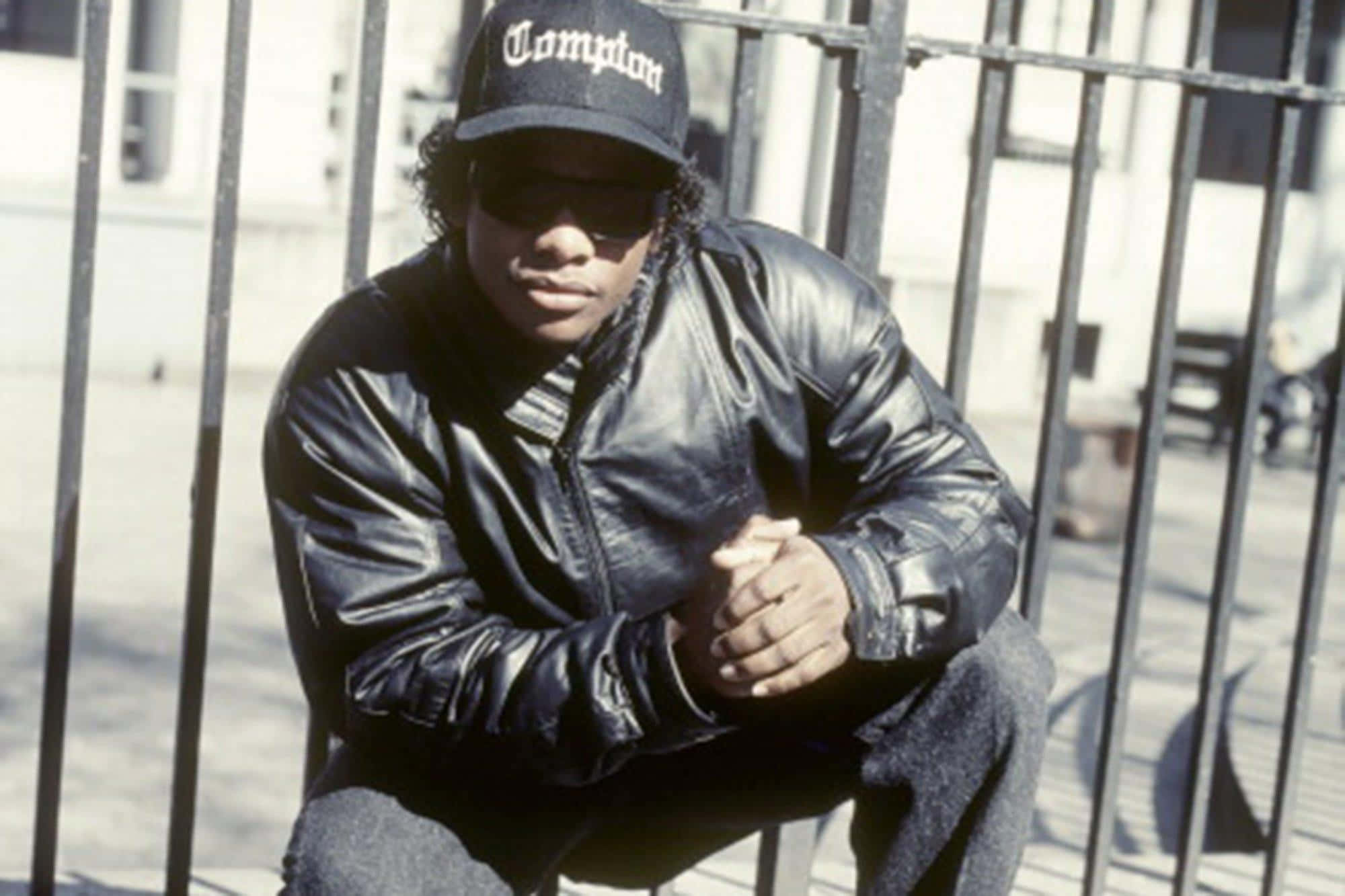 Eazy E 桌布 壁紙 — 一位戴著Compton帽和皮褸的年輕男子自信地坐在金屬圍欄上，在明亮的日光下。