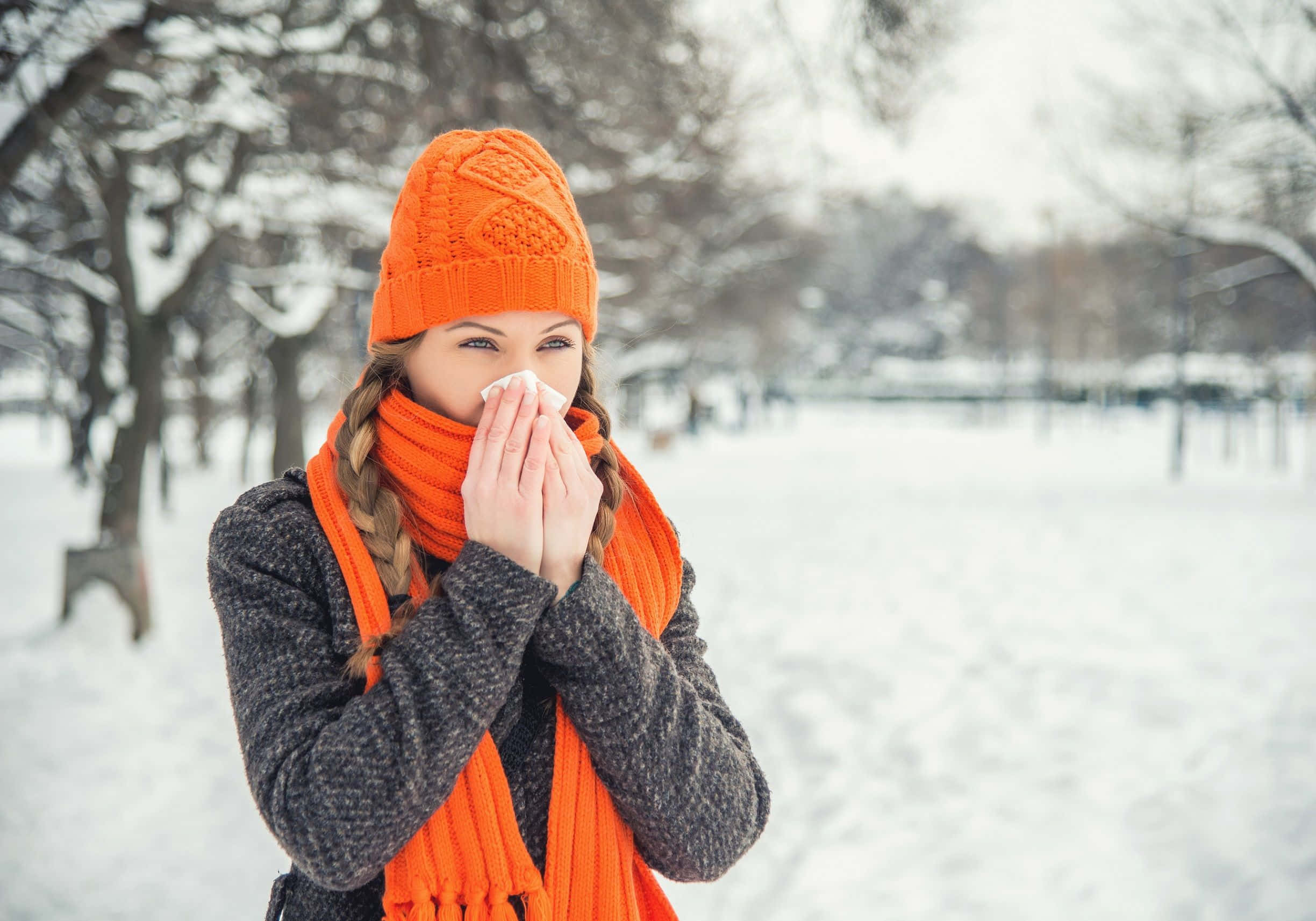 Cold 桌布 壁紙 — 一位戴著亮橙色針織帽和圍巾的女性，在白雪覆蓋的公園裡對著紙巾打噴嚏。