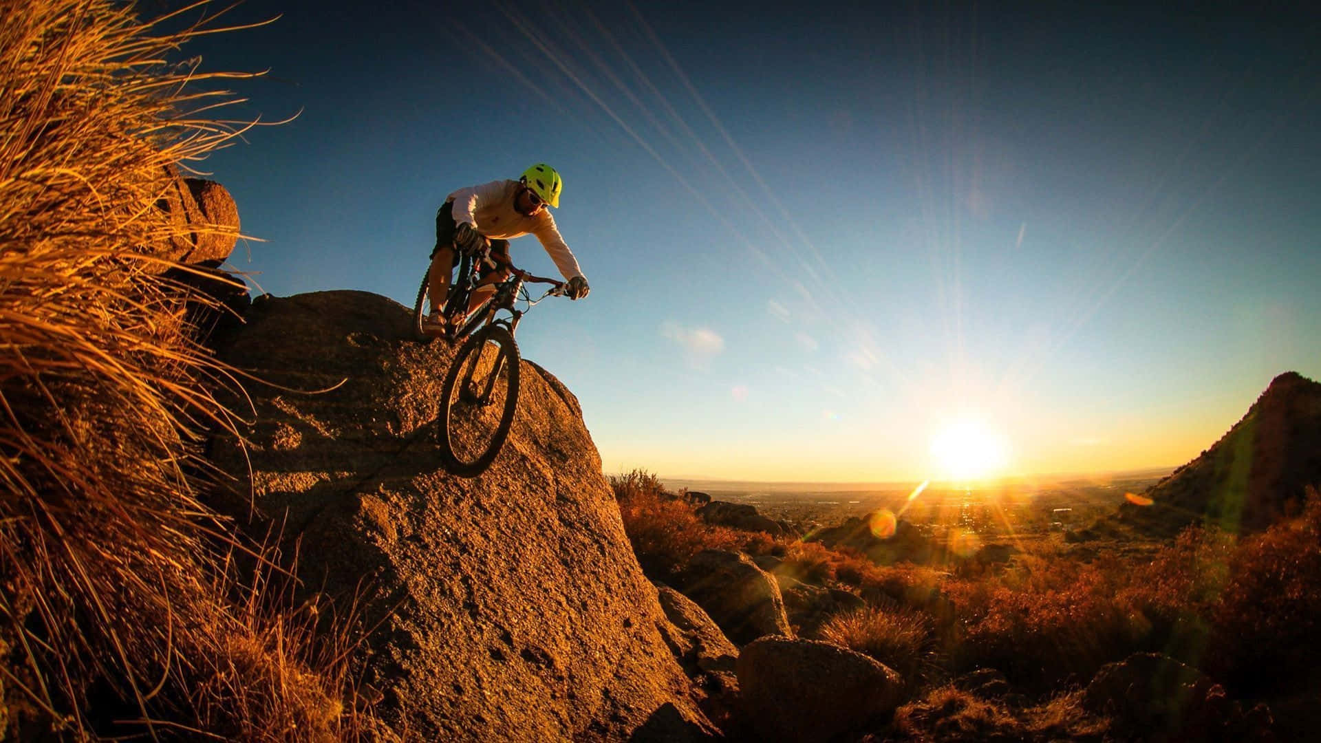 Mtb 桌布 壁紙 — 一位戴著亮色頭盔的山地車手在岩石地形上騎行，夕陽在遠山後落下，灑下金色光芒。