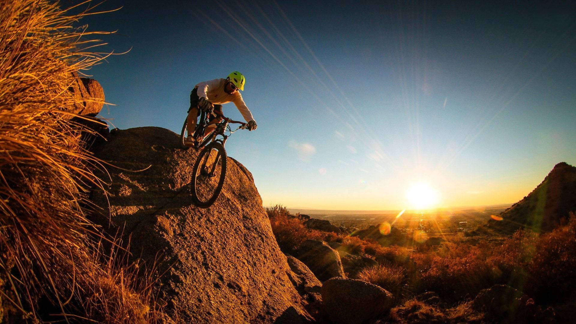 Mountain Bike 桌布 壁紙 — 一位戴著亮色頭盔的山地車手在岩石小徑上騎行，夕陽在遠山後落下，灑下金色光芒。