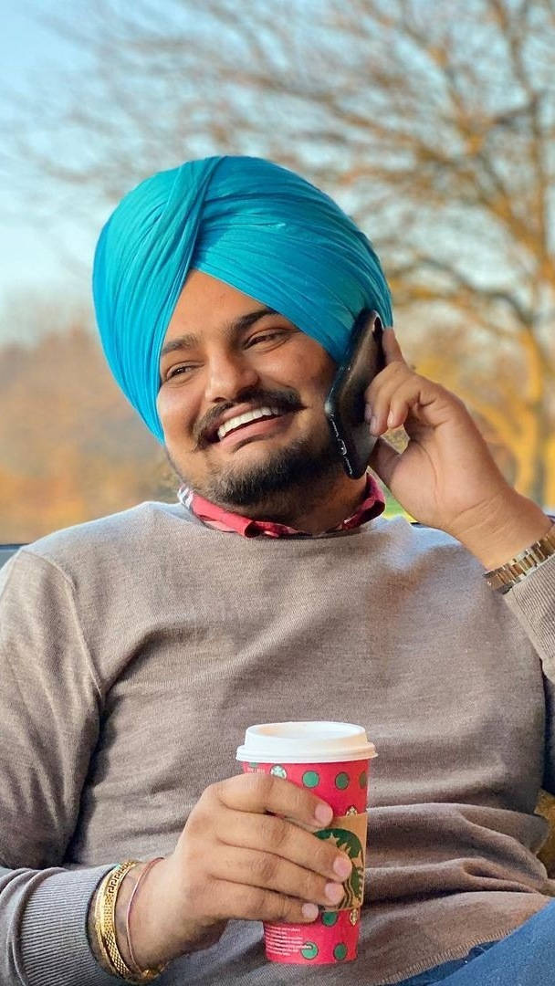 Indian 桌布 壁紙 — 一位戴著亮藍色頭巾的微笑男子，在戶外一邊拿著色彩繽紛的咖啡杯，一邊講電話。