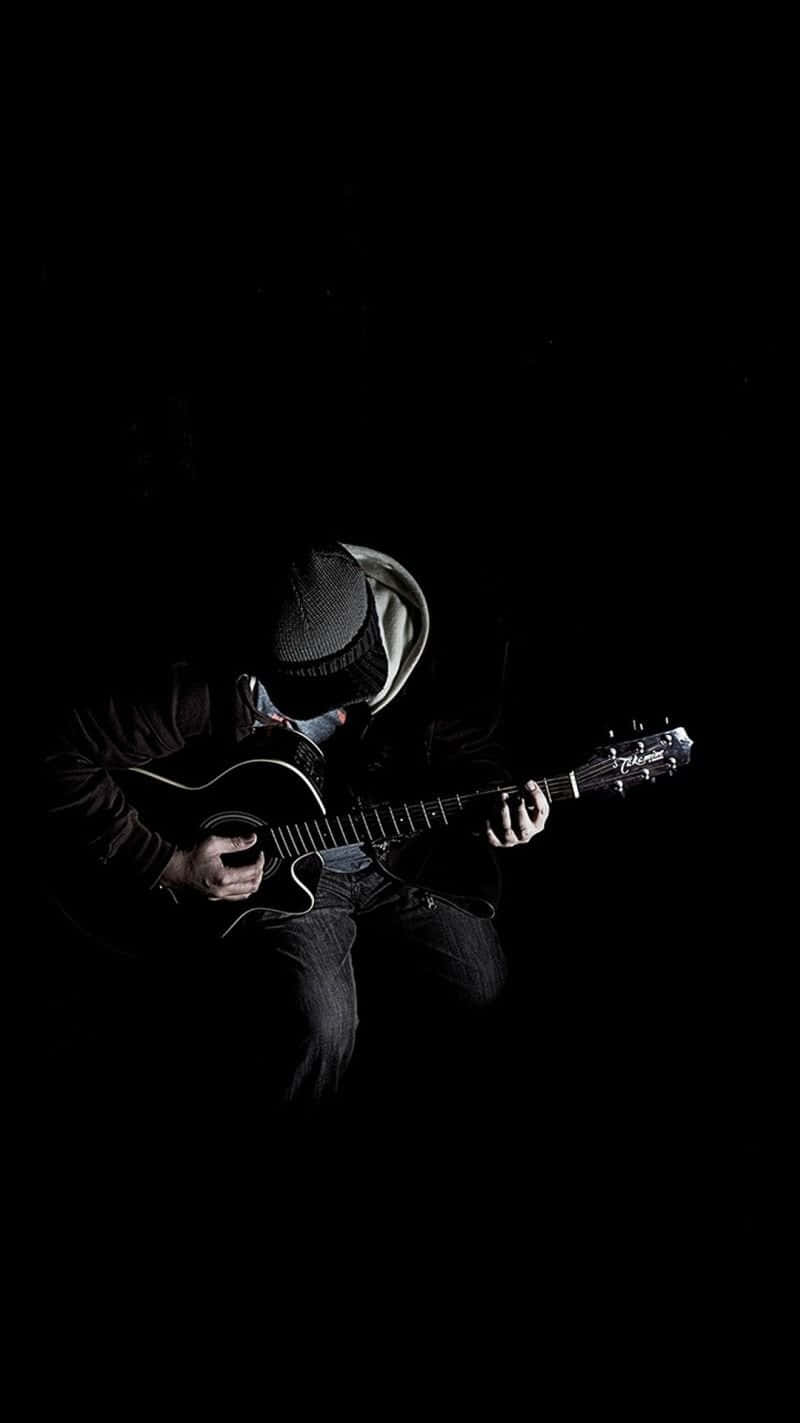 Musical 桌布 壁紙 — 一位戴著冷帽的音樂家在戲劇性的聚光燈下，在漆黑背景前彈奏木結他。