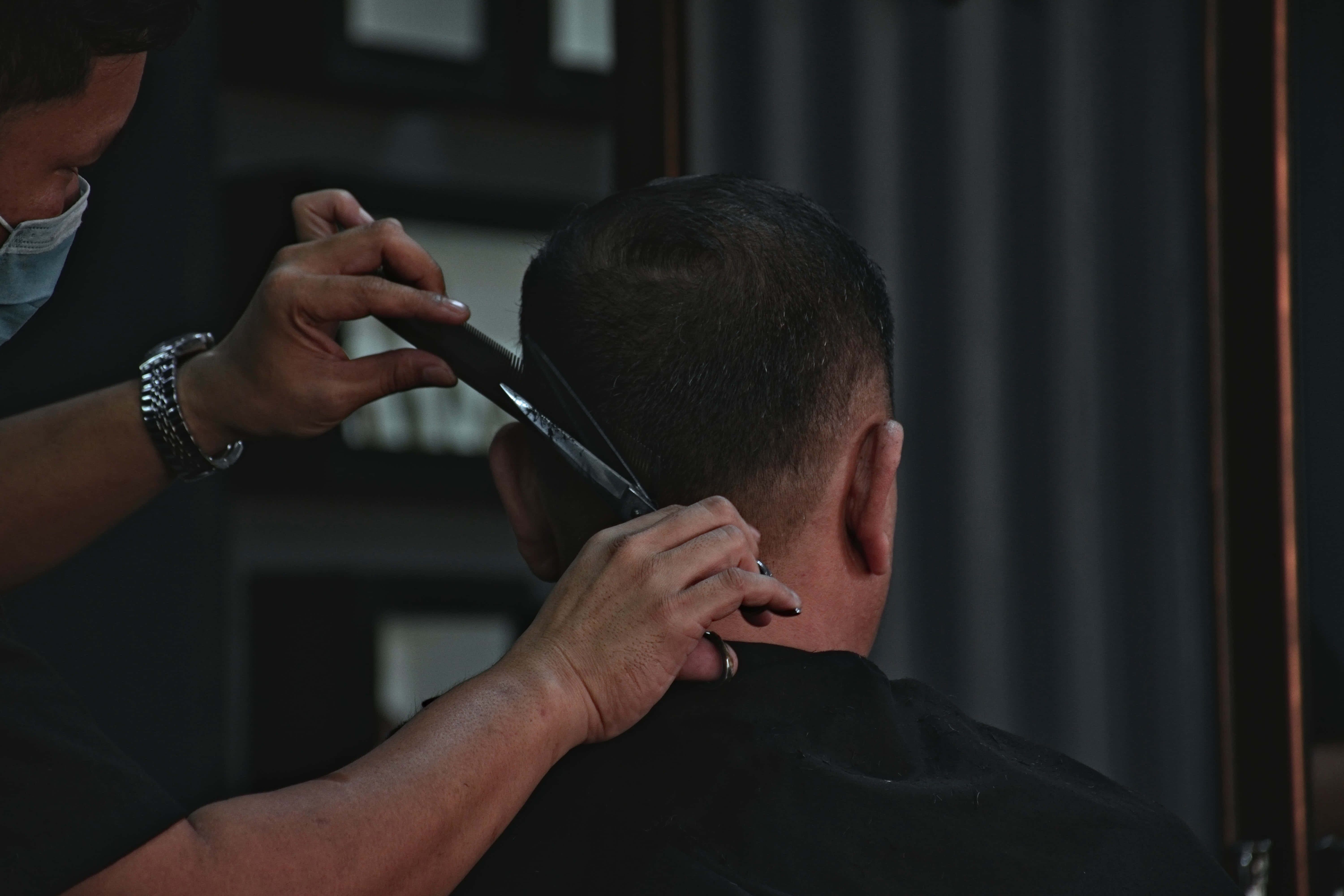 Barber 桌布 壁紙 — 一位戴著口罩的理髮師在燈光昏暗的沙龍裡，用剪刀仔細修剪一位男士的短黑髮。