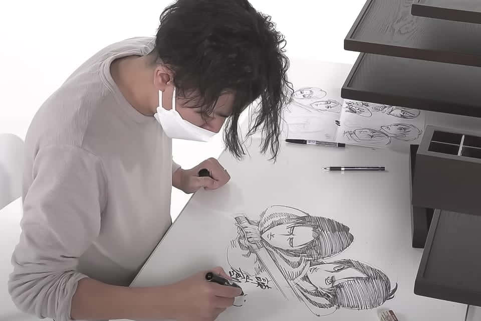 Isayama Hajime 桌布 壁紙 — 一位戴著口罩的藝術家俯身在一張白色桌子上，用黑色馬克筆專注地素描動漫角色。