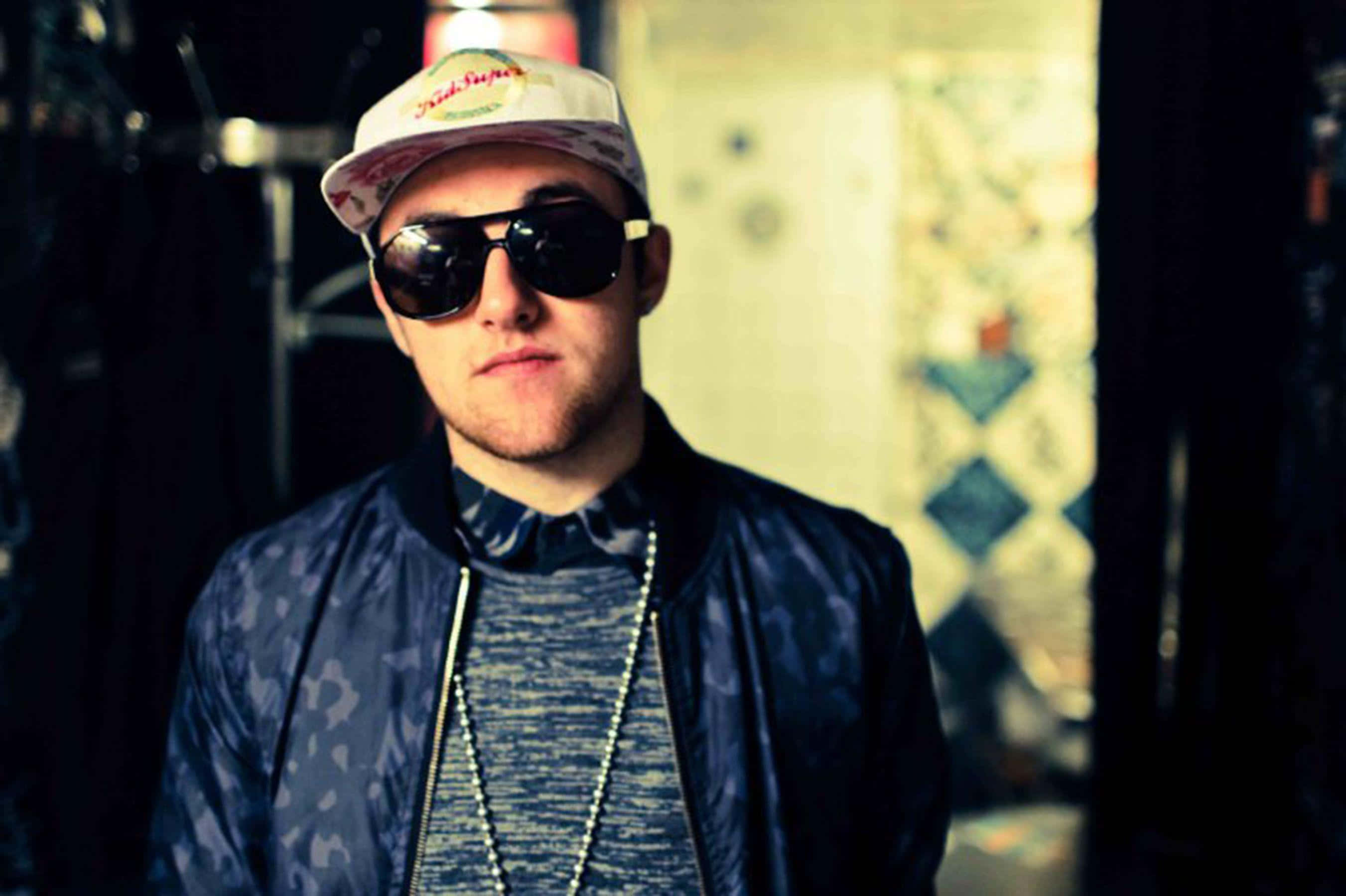 Mac Miller 桌布 壁紙 — 一位戴著圖案帽和太陽眼鏡的年輕男子，在昏暗的城市小巷中自信地凝視鏡頭。