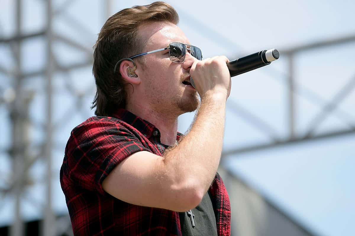 Morgan Wallen 桌布 壁紙 — 一位戴著太陽眼鏡和紅色格仔恤衫的男歌手，在明亮的日光下於戶外音樂會中熱情地對著咪高峰演唱。