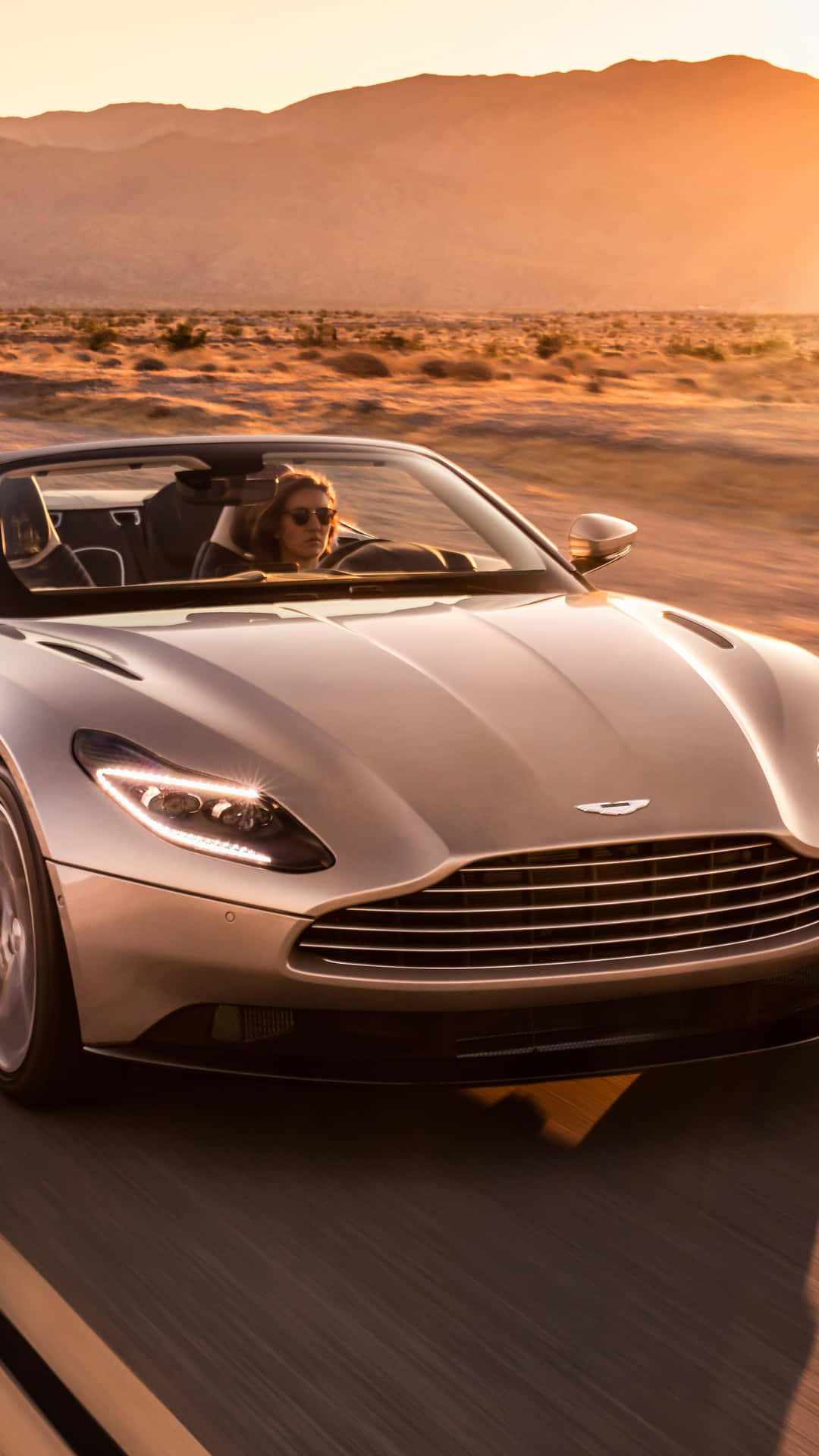 Aston Martin Db11 桌布 壁紙 — 一位戴著太陽眼鏡的女司機駕駛銀色Aston Martin DB11開篷跑車在日落時分疾馳穿過沙漠。