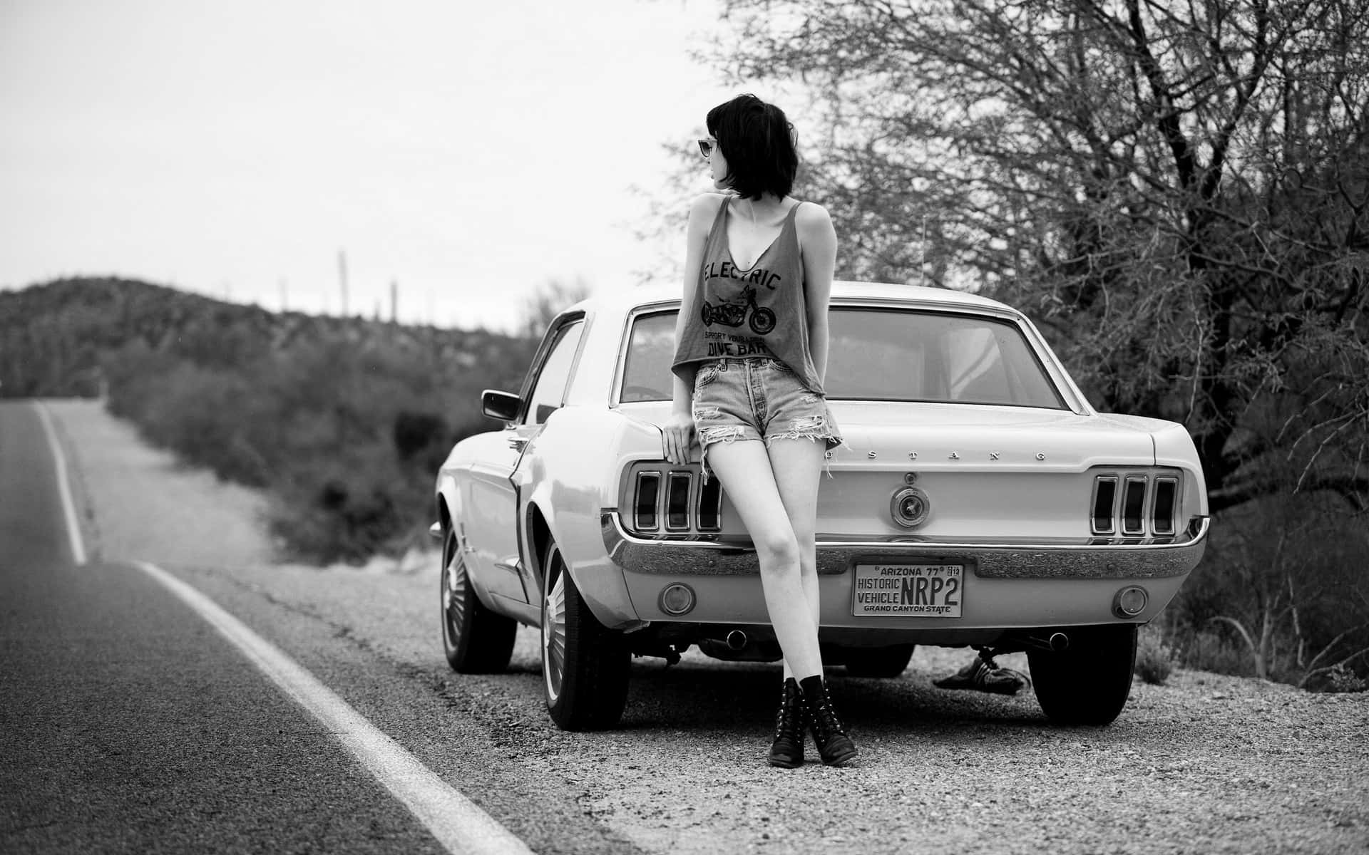 Black And White Vintage Car 桌布 壁紙 — 一位戴著太陽眼鏡的女性在沙漠路邊靠在一輛復古野馬車上，以光禿的樹木和遠山為框架，呈現黑白風格。
