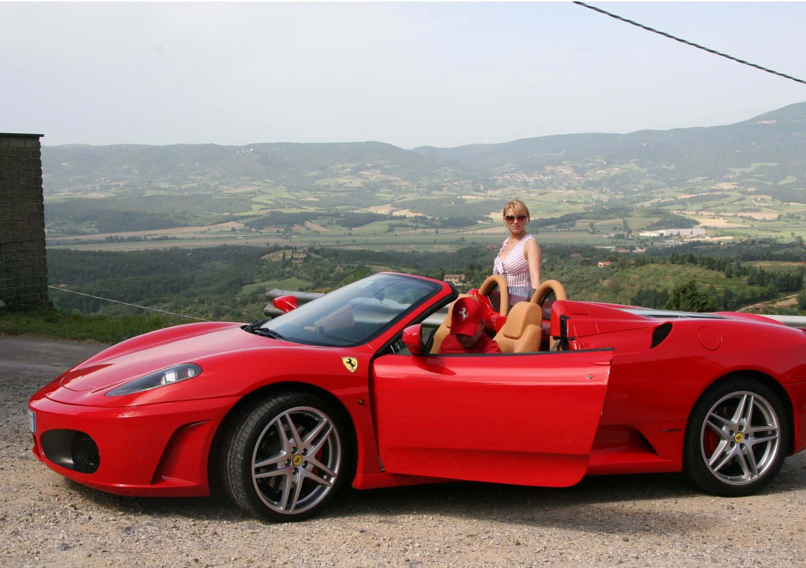 Ferrari F430 桌布 壁紙 — 一位戴著太陽眼鏡的女性站在一輛鮮紅色法拉利開篷車旁，車停在碎石觀景點上，眼前是起伏的綠色山丘和遠處村莊，天空朦朧。