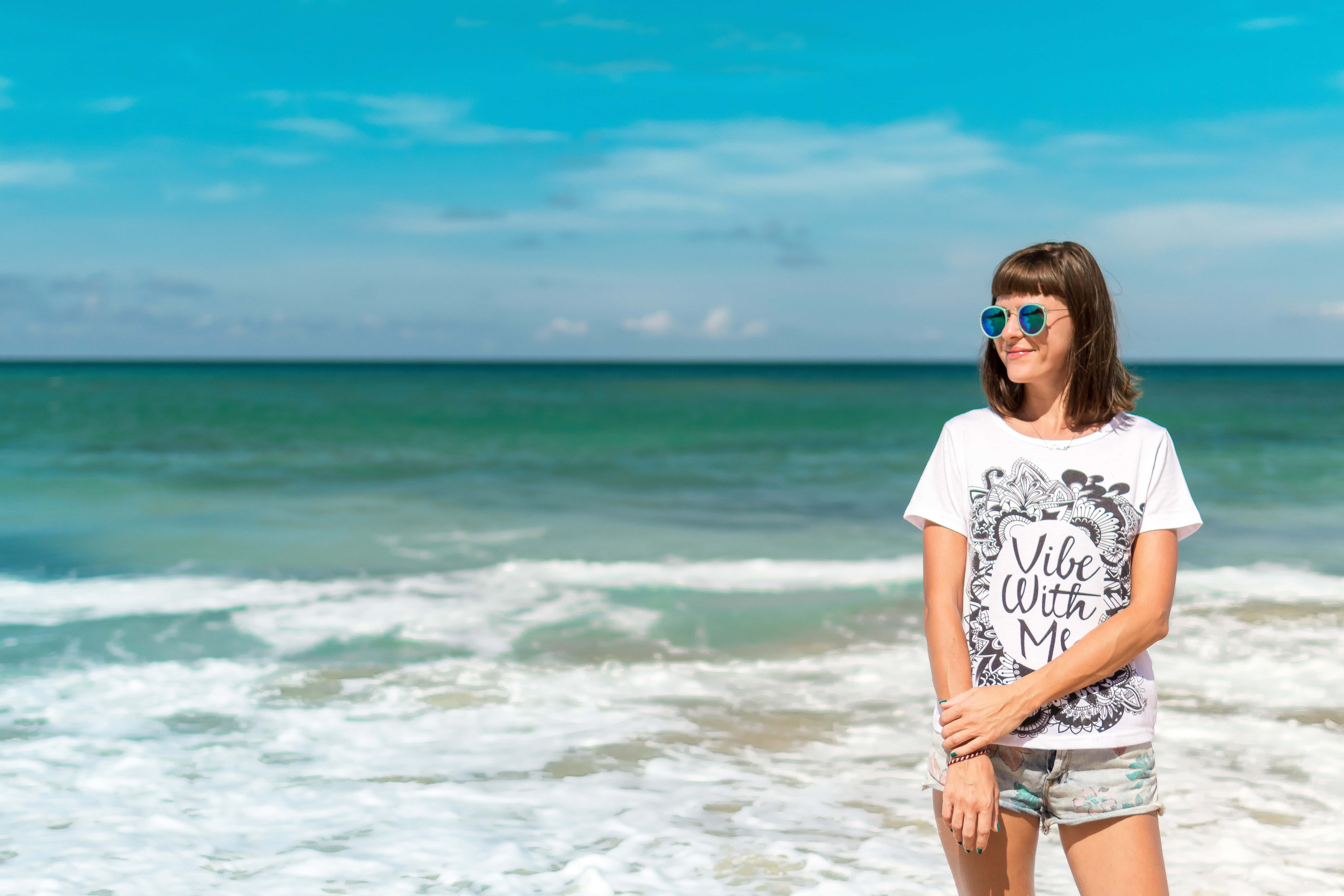 Summer Vacation 桌布 壁紙 — 一位戴著太陽眼鏡的微笑女士站在海邊，海浪在明亮的藍天下拍打。