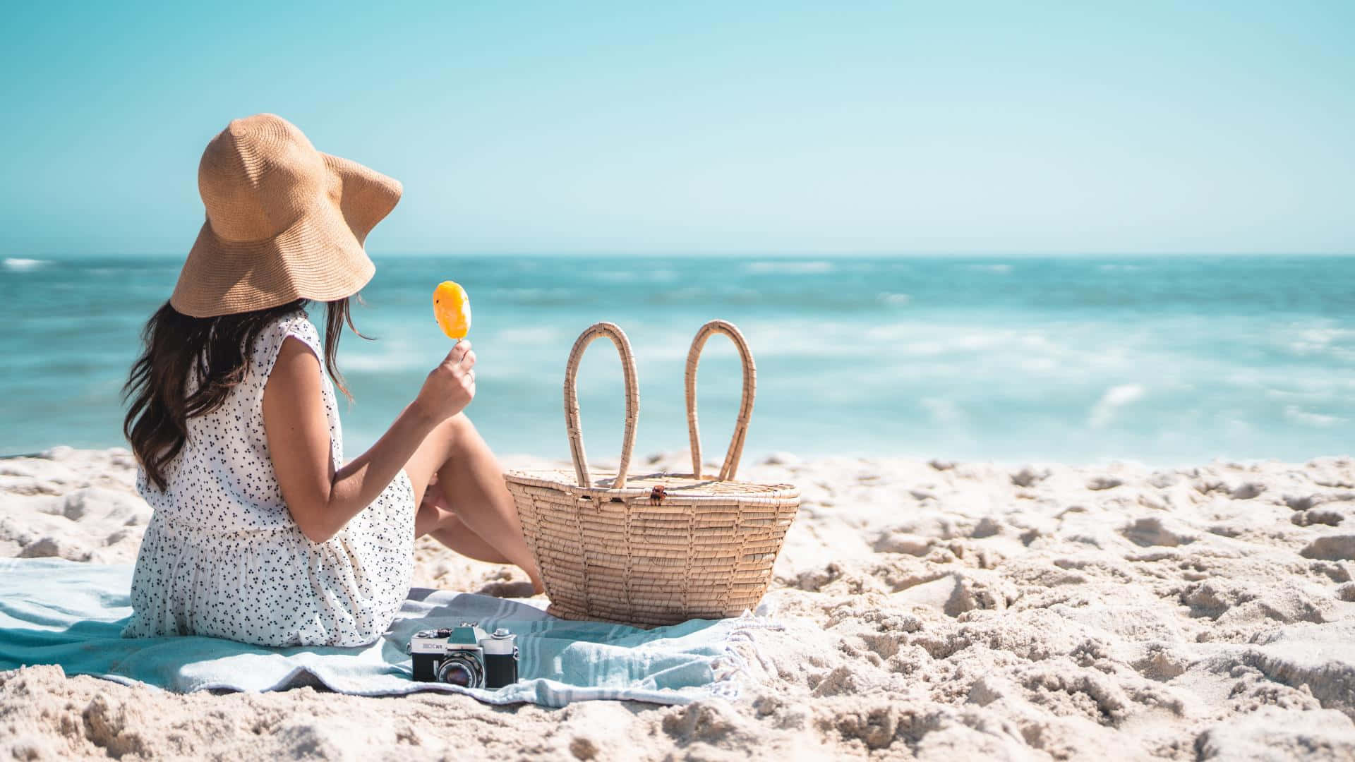 Beach Picnic 桌布 壁紙 — 一位戴著寬邊帽的女性在陽光明媚的海灘上，在柳條籃和復古相機旁邊享用冰棒。