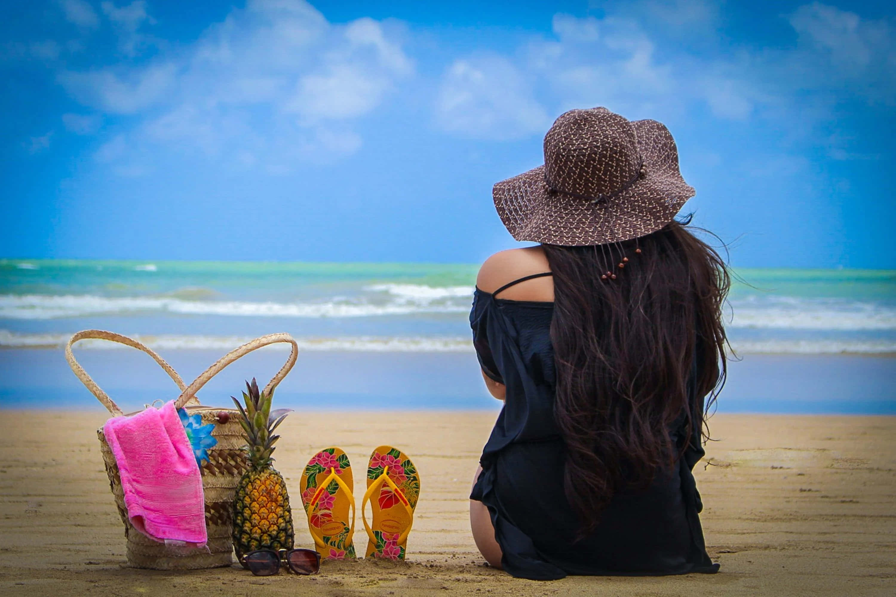 Beach Bag 桌布 壁紙 — 一位戴著寬邊帽的女性坐在沙灘上，面對著在明亮藍天和蓬鬆雲朵下的碧綠海浪。