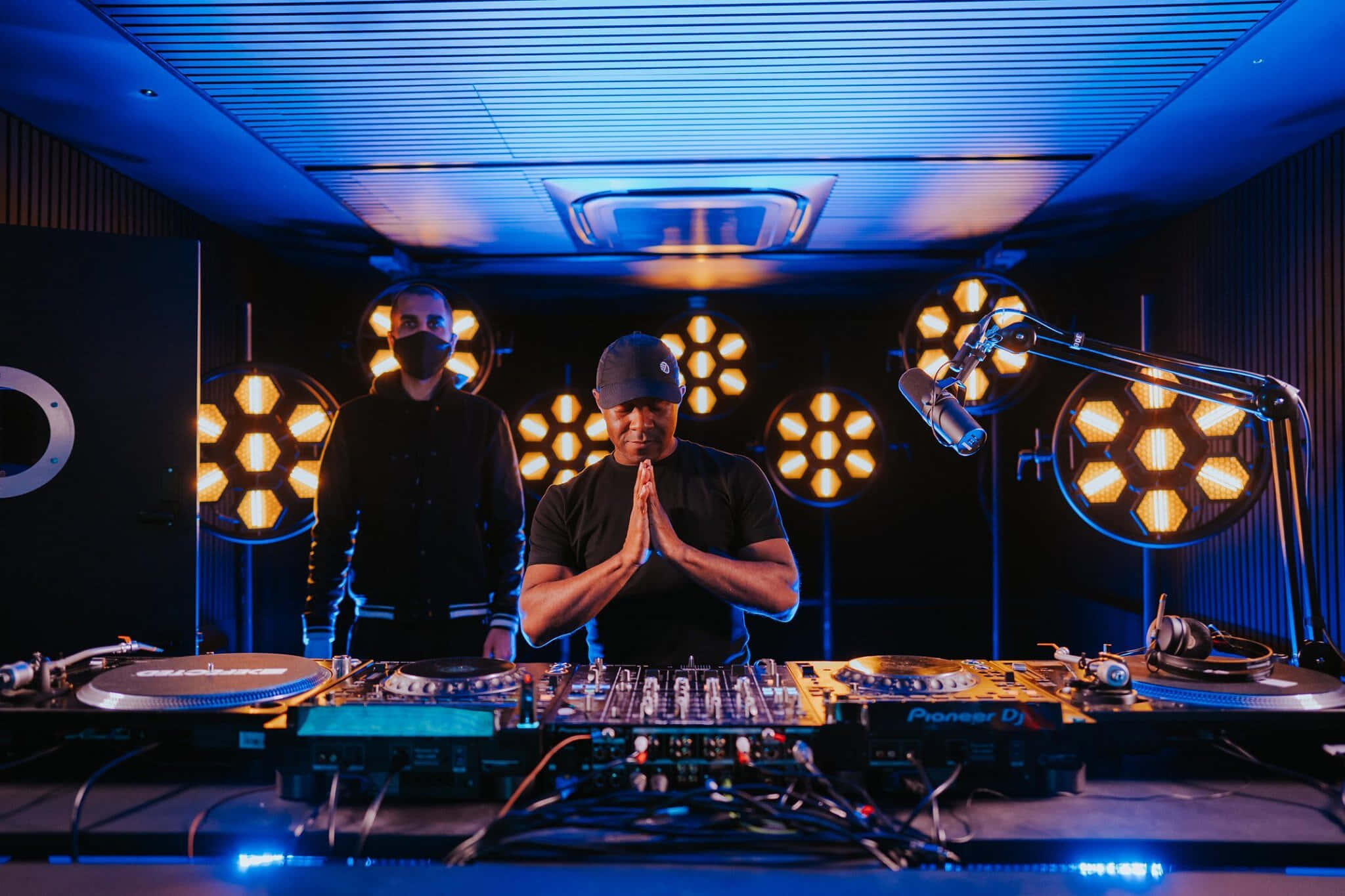DJ 桌布 壁紙 — 一位戴著帽子的DJ站在Pioneer DJ設備後面，雙手緊握，下方是發光的六角形燈光和藍色環境照明。