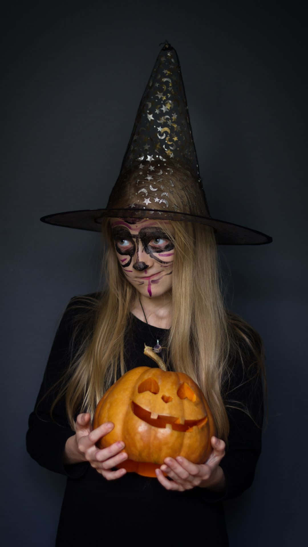 Horror Costumes 桌布 壁紙 — 一位戴著星星帽和骷髏妝容的年輕女巫，在黑暗的攝影棚背景下手持雕刻的傑克南瓜燈。