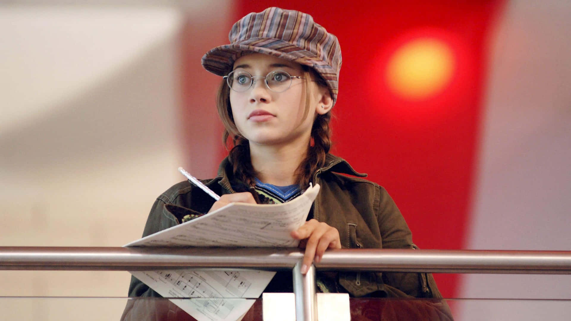 High School Musical 桌布 壁紙 — 一位戴著條紋帽和眼鏡的年輕女性，靠在紅色牆壁的欄杆上，若有所思地拿著文件。