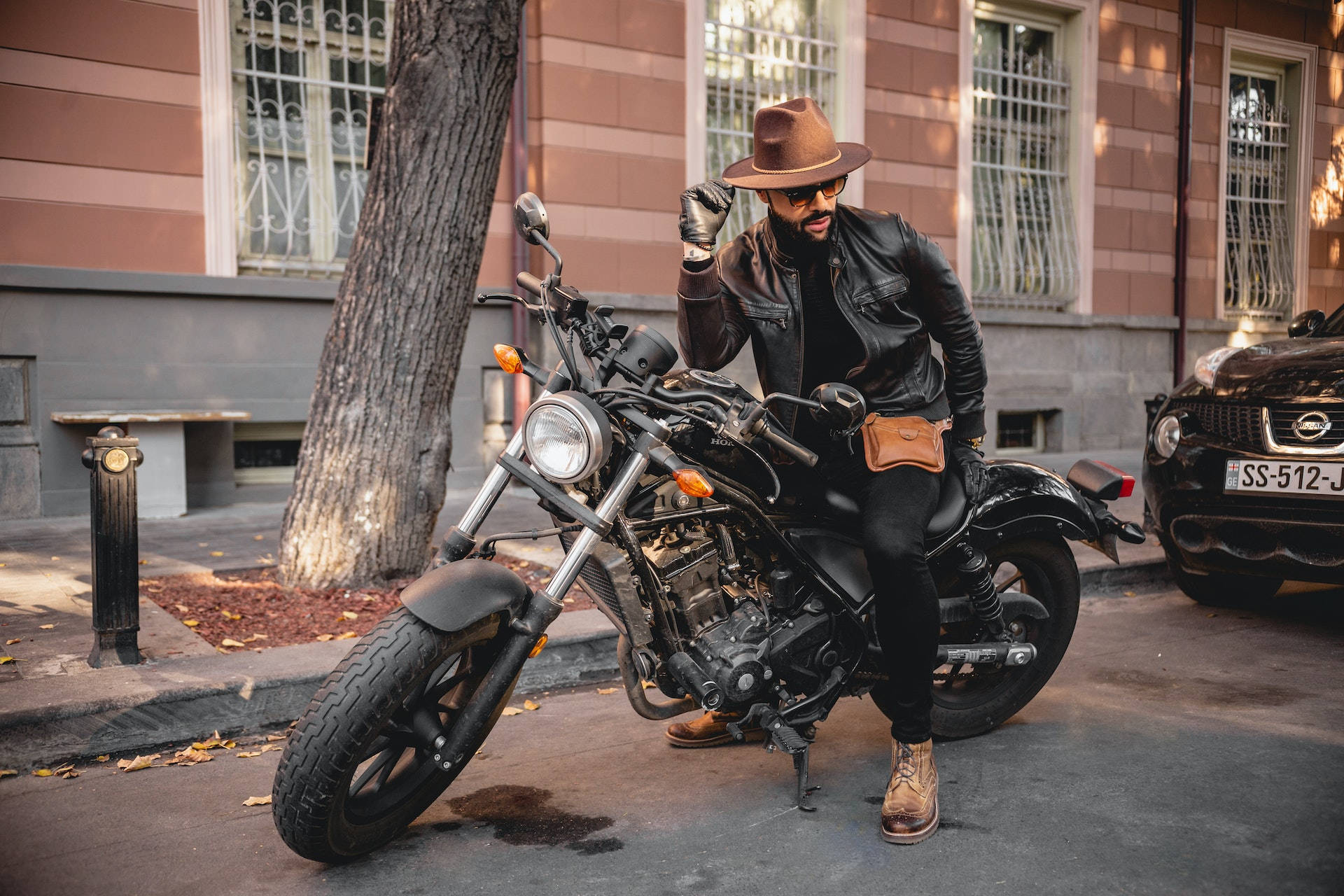 Bike Rider 桌布 壁紙 — 一位戴著棕色帽子和皮褸的時尚男士，自信地坐在一輛停泊在城市行人路上的黑色巡航電單車上，旁邊有一棵樹和一座復古建築。