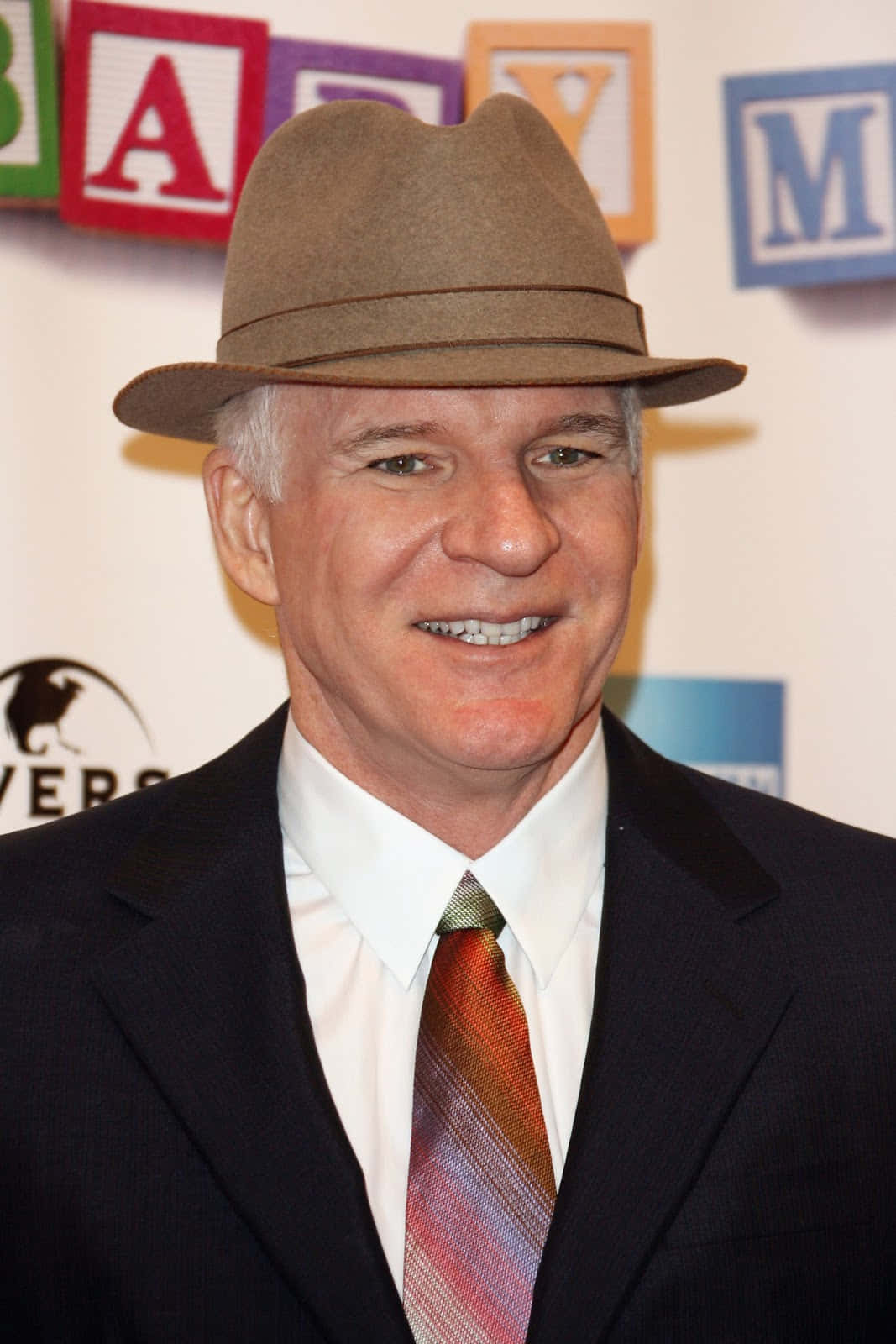 steve martin 桌布 壁紙 — 一位戴著棕色軟呢帽和深色西裝的微笑男子，在活動中站在色彩繽紛的文字背景前。
