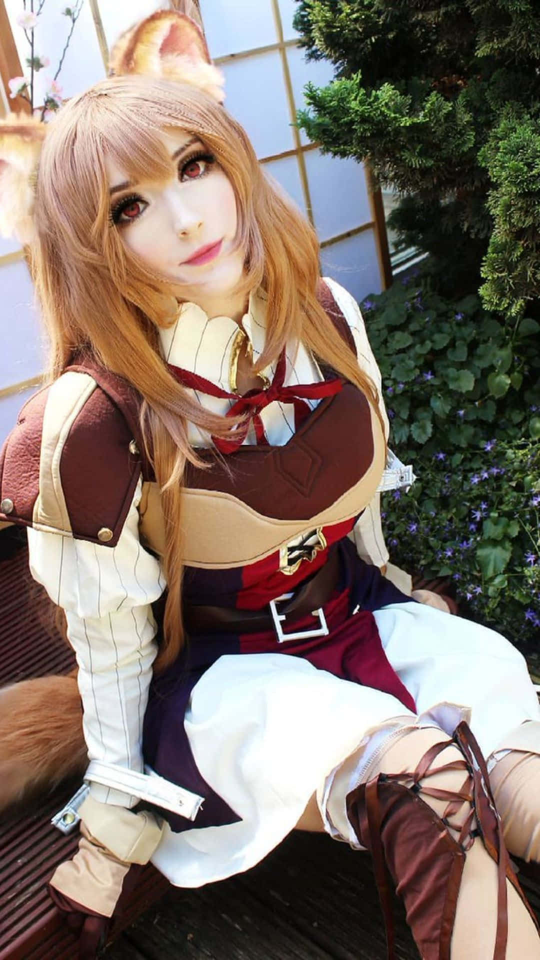 Cosplay Anime Kawaii 桌布 壁紙 — 一位戴著狐狸耳朵和尾巴的cosplayer，穿著精緻的奇幻服裝，坐在戶外，背景是綠植和木柵欄。