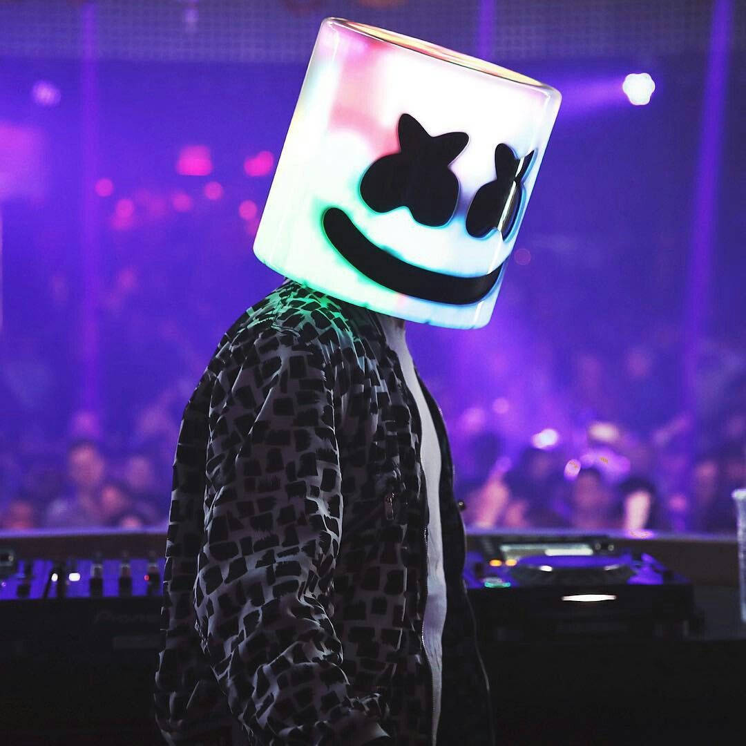marshmello iPhone 桌布 壁紙 — 一位戴著發光立方體面具的DJ站在混音台前，舞台紫色燈光下，背後是模糊的人群。