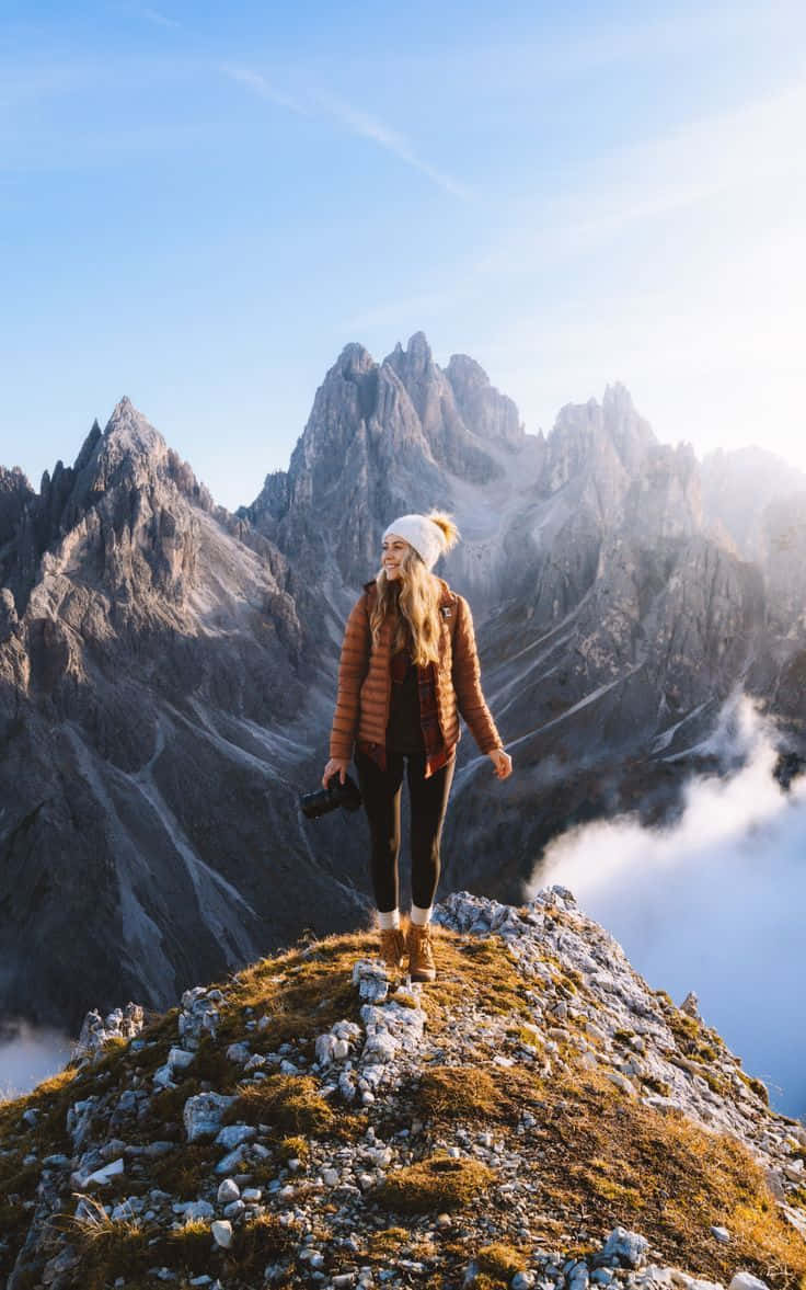 Winter Hiking 桌布 壁紙 — 一位戴著白色冷帽和棕色外套的女性站在岩石山頂上微笑，雲朵在她下方飄過，襯著清澈的藍天。