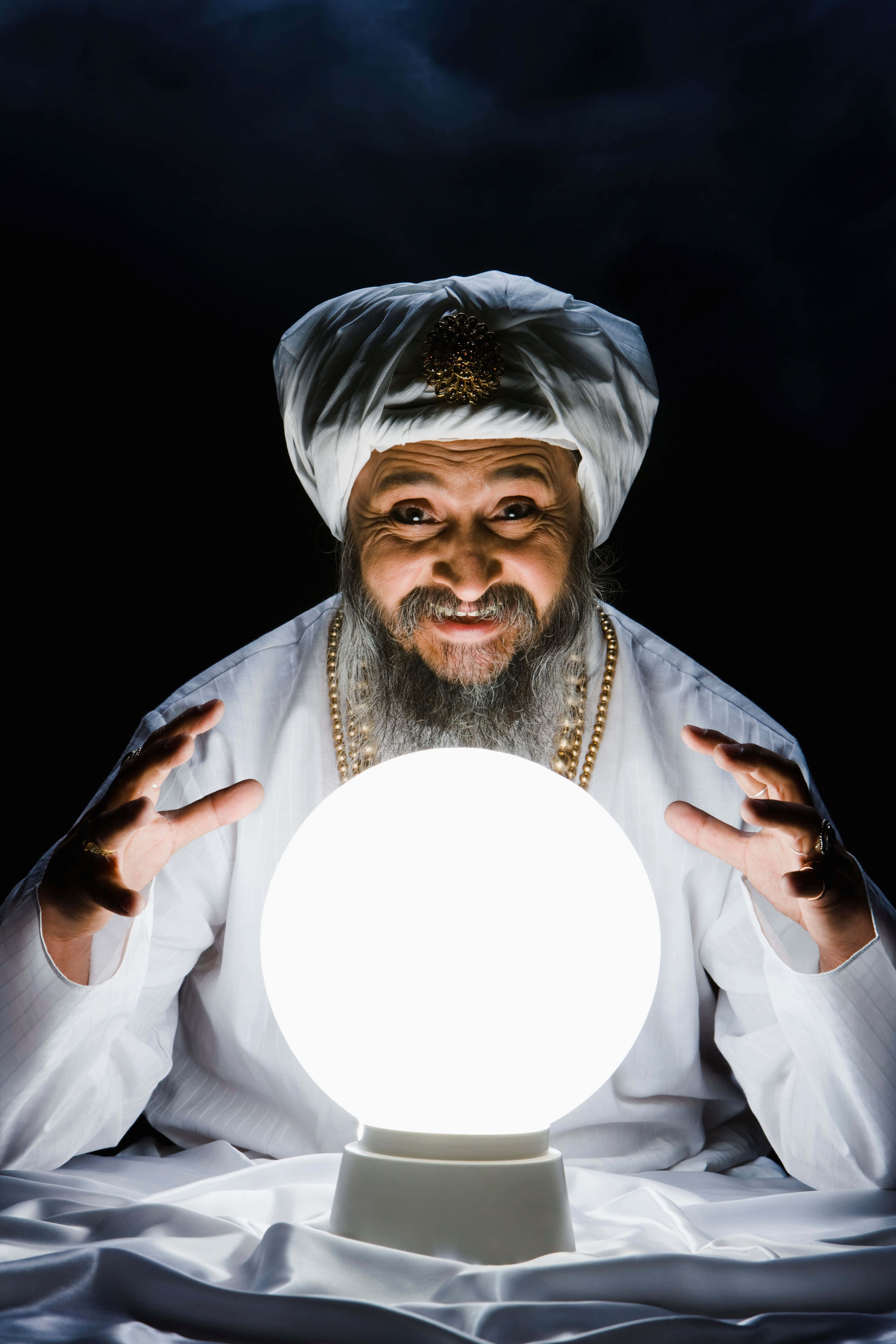 Fortune Teller 桌布 壁紙 — 一位戴著白色頭巾、留著鬍子的年長神秘主義者，在黑暗背景下專注地凝視著一顆發光的水晶球。