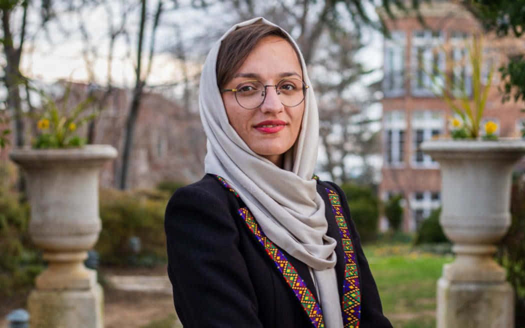 Taliban 桌布 壁紙 — 一位戴著米色頭巾和眼鏡的女性在花園中微笑，身後有石甕和模糊的磚建築。