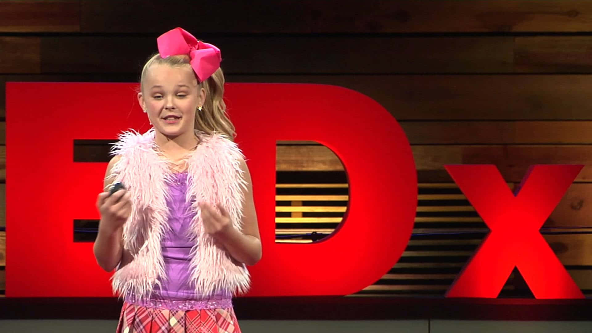 jojo siwa 桌布 壁紙 — 一位戴著粉紅色蝴蝶結和羽毛背心的年輕女孩在身後有紅色字母的TEDx舞台上熱情地演講。