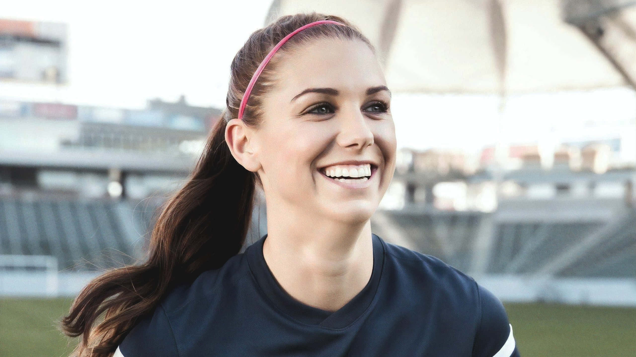 Alex Morgan 桌布 壁紙 — 一位戴著粉紅色頭帶的快樂年輕女子在體育場內穿著深色運動衫燦爛地微笑。