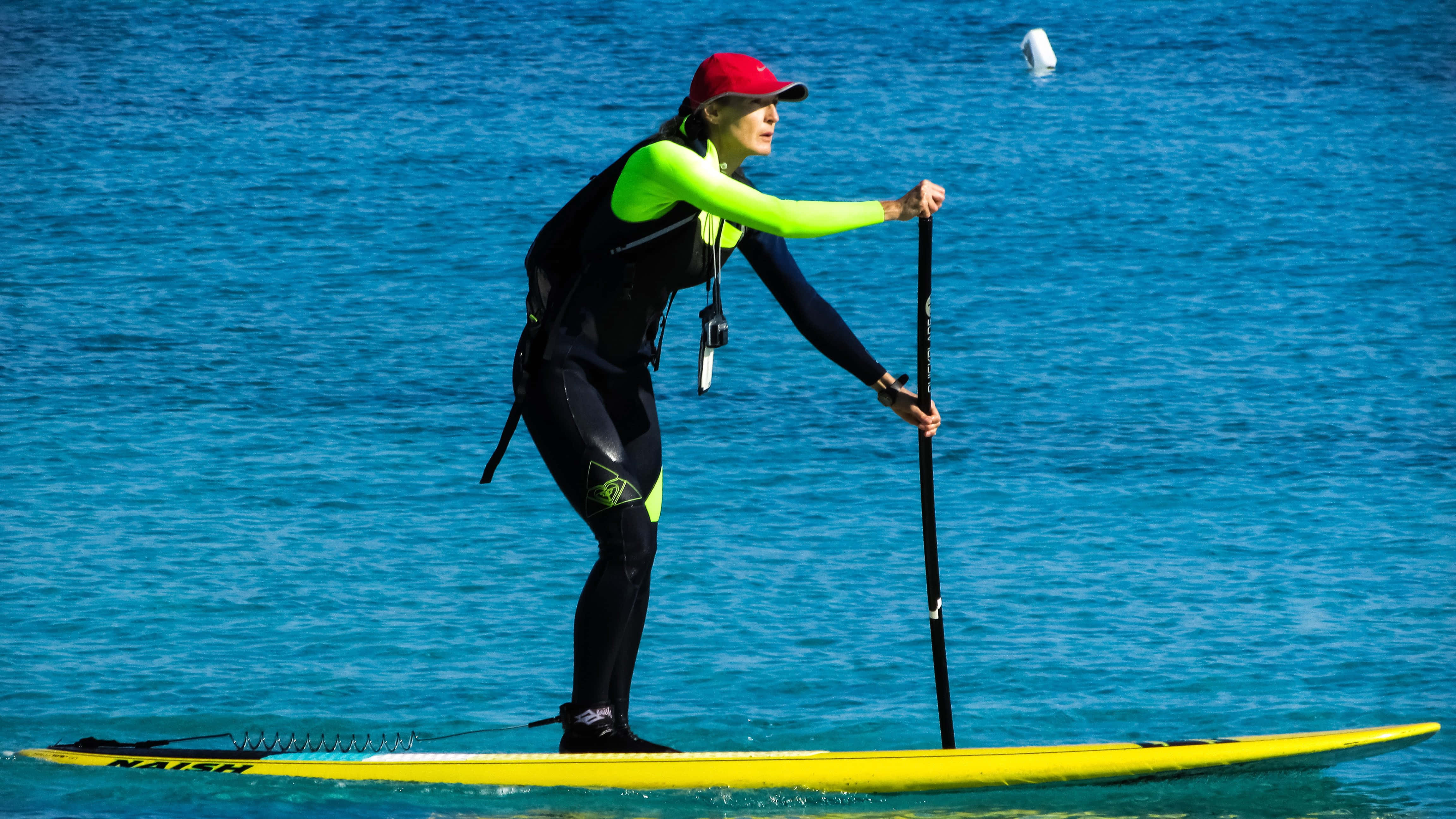 Paddleboarding 桌布 壁紙 — 一位戴著紅色帽子和螢光潛水衣的女子，在平靜的碧綠水面上划著亮黃色的直立槳板。