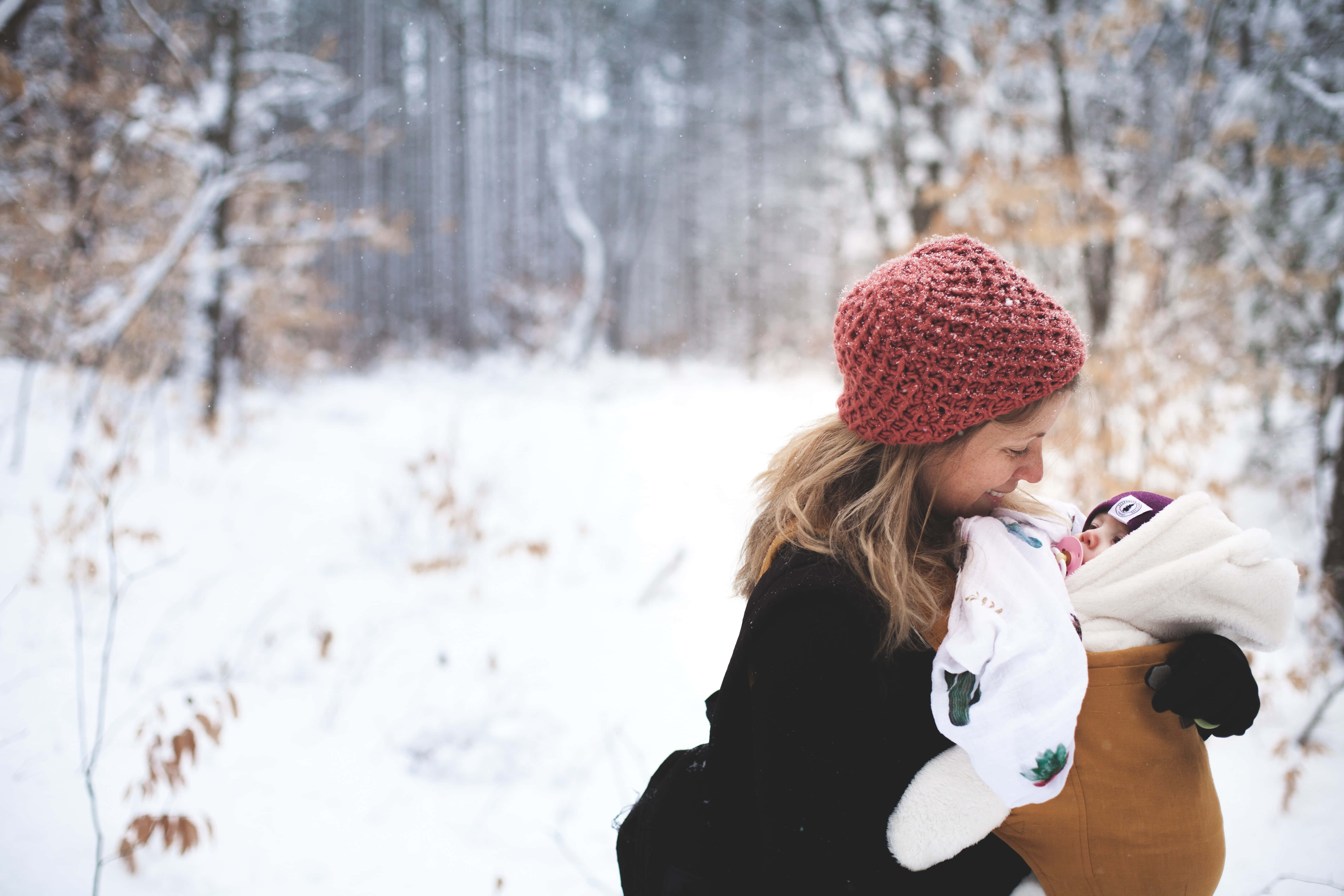 Mom 桌布 壁紙 — 一位戴著紅色針織帽子的女士在雪地森林中溫柔地抱著一個裹著白色雪地裝備的嬰兒。