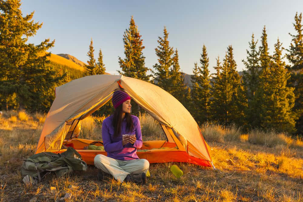 Fall Camping 桌布 壁紙 — 一位戴著紫色冷帽的女性盤腿坐在橙色帳篷外，啜飲咖啡，金色夕陽灑在松樹和山巒上。